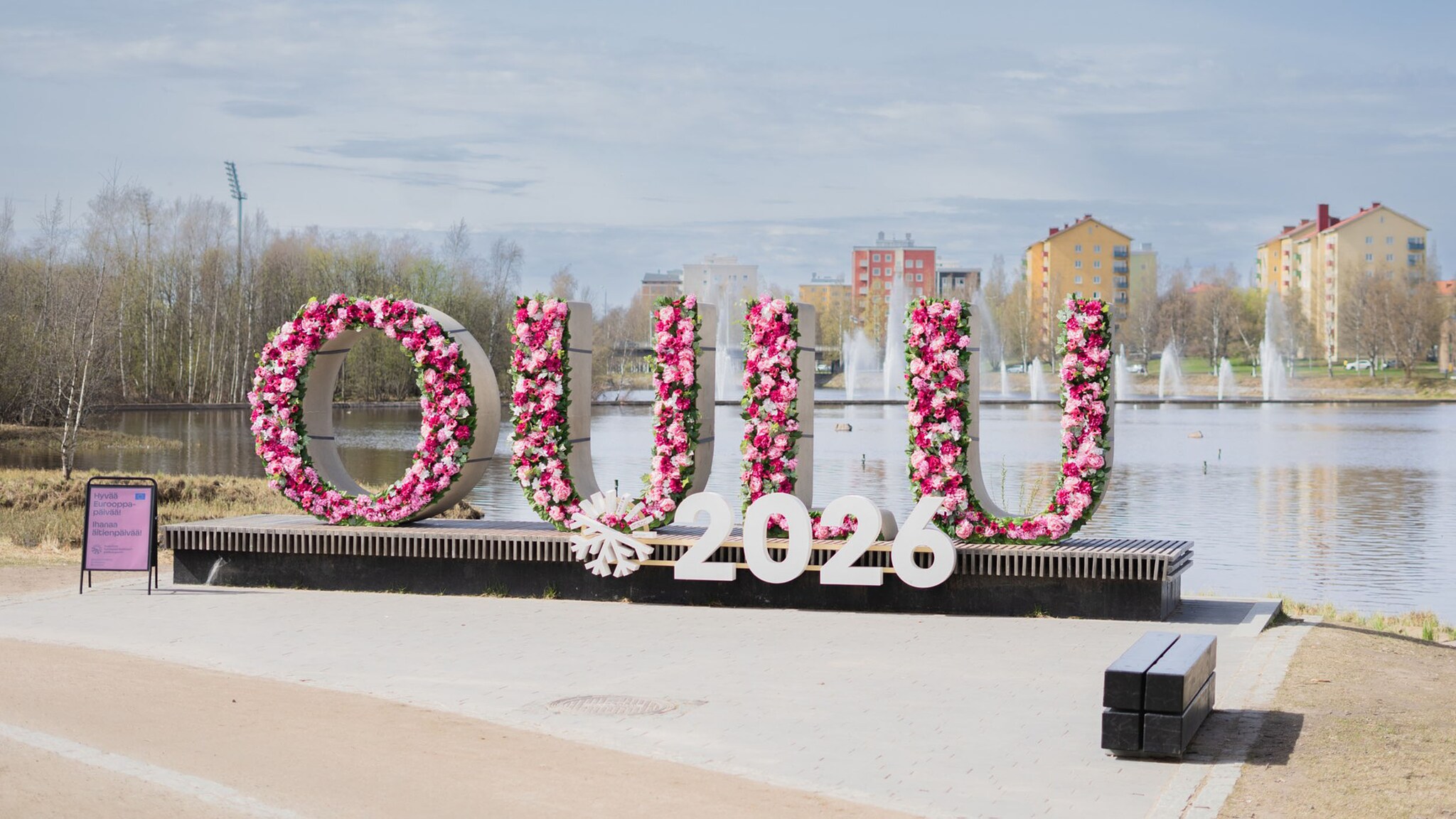Ein großer, mit pinken Blumen geschmückter Schriftzug mit den Buchstaben OULU steht vor einem See in einer Stadt. Davor ein Logo mit der Zahl 2026.