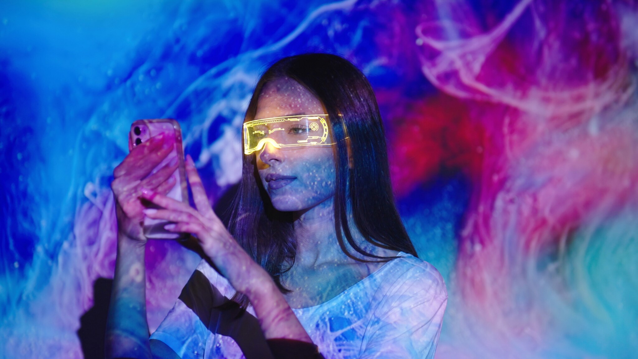 Eine Frau mit gelb leuchtender Cyberpunk LED-Brille schaut auf ein Smartphone in ihren Händen vor einer Wand, angeleuchtet von einer Lichtprojektion mit blau-pinkem Muster.