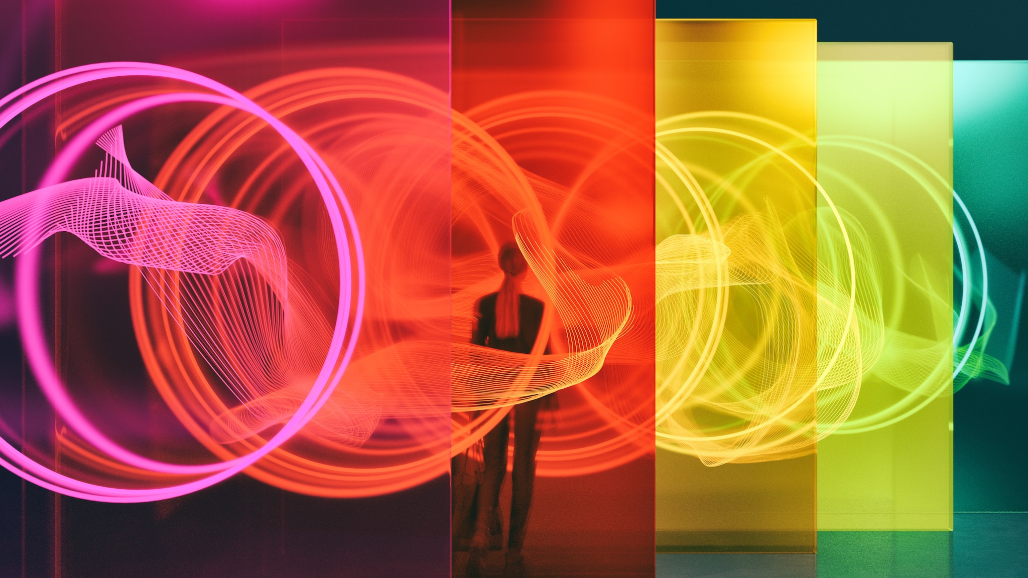 amex-magazin-digitale-kunst-transparenten-waende Rückansicht einer Frau, die zwischen transparenten Wänden in unterschiedlichen Farben mit runden Lichtstreifen steht in einer Halle.