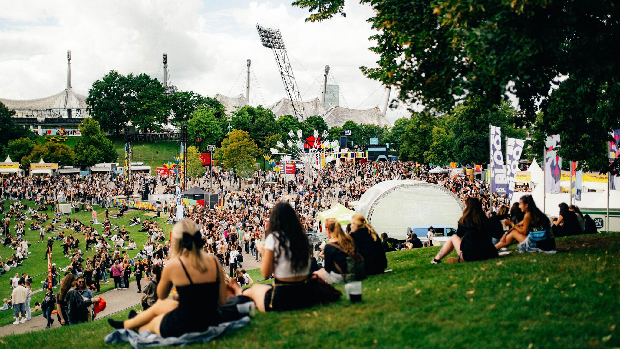 Zahlreiche Menschen stehen und sitzen auf grünen Hügeln im Olympiapark während eines Festivals im Sommer..