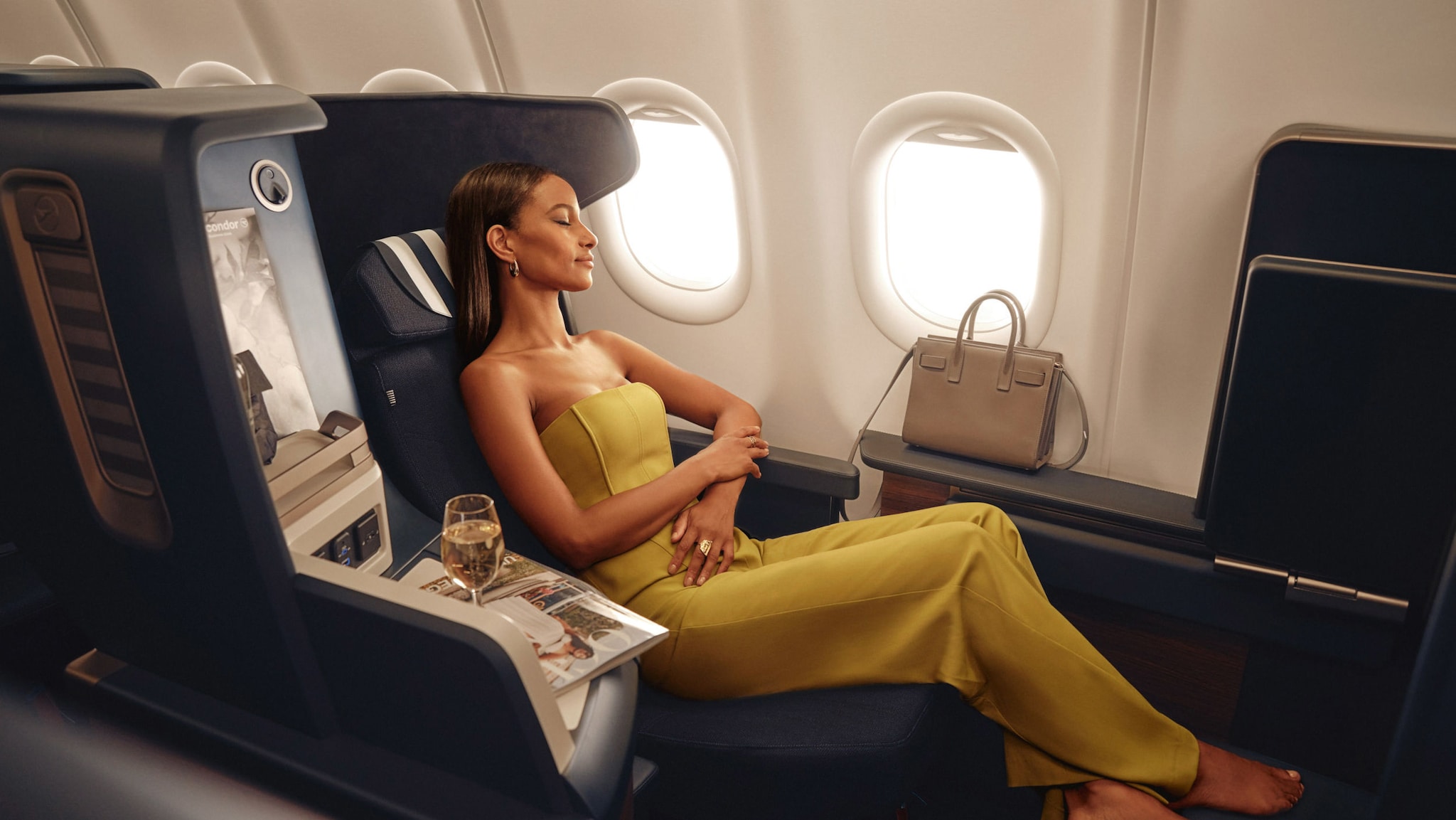 Eine Frau in einem eleganten, gelben Kleid sitzt mit geschlossenen Augen und ausgestreckten Beinen auf einem luxuriösen Fensterplatz der Business Class in einem Flugzeug, neben ihr befinden sich Zeitschriften und ein Glas Champagner auf einer Ablage
