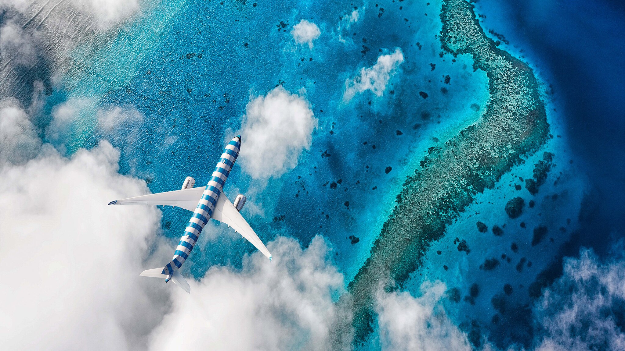Aufsicht eines blau-weiß gestreiften Flugzeuges zwischen Wolken in der Luft über einem Korallenriff im türkisblauen Meer.