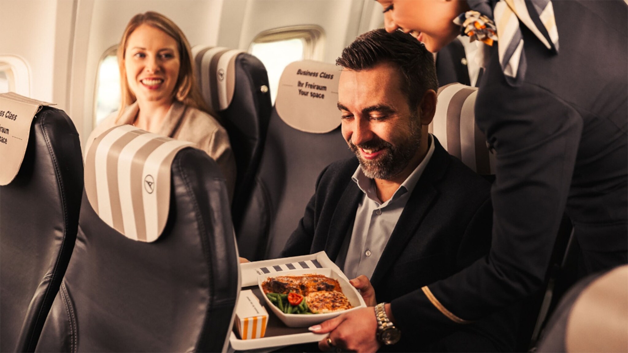Eine Stewardess serviert einem lächelnden Flugzeugpassagier eine Mahlzeit auf einem Tablett, eine Frau in derselben Sitzreihe am Fensterplatz schaut ihnen zu.