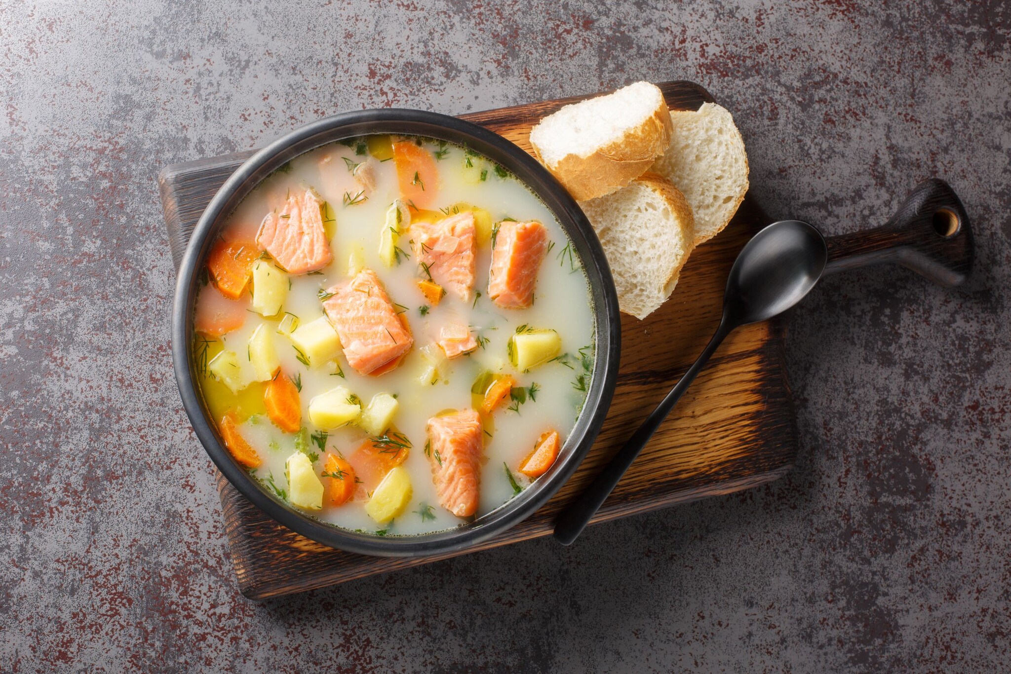 Blick von oben auf eine Schüssel mit Lachsfischsuppe, die mit Brotscheiben und einem Löffel auf einem Holzbrett serviert wird. 