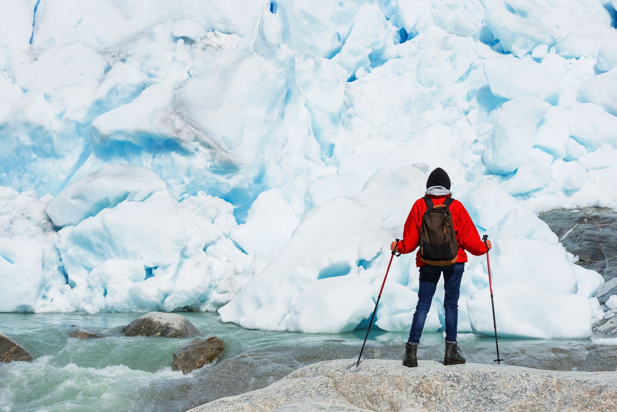 Rückansicht einer Person mit Wanderstöcken vor einer Eiswand am Nigardsbreen. 