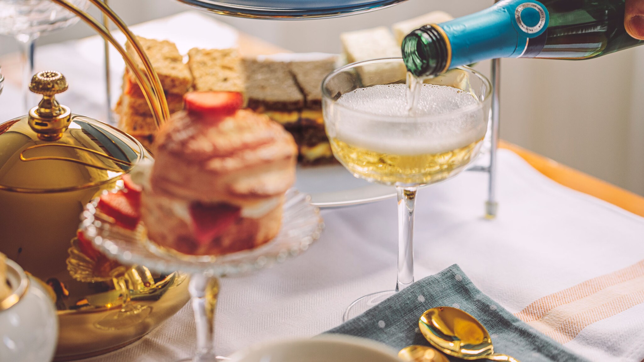 Heller Schaumwein wird aus einer Flasche in eine Sektschale auf einem elegant eingedeckten Tisch mit süßem Gebäck eingefüllt.