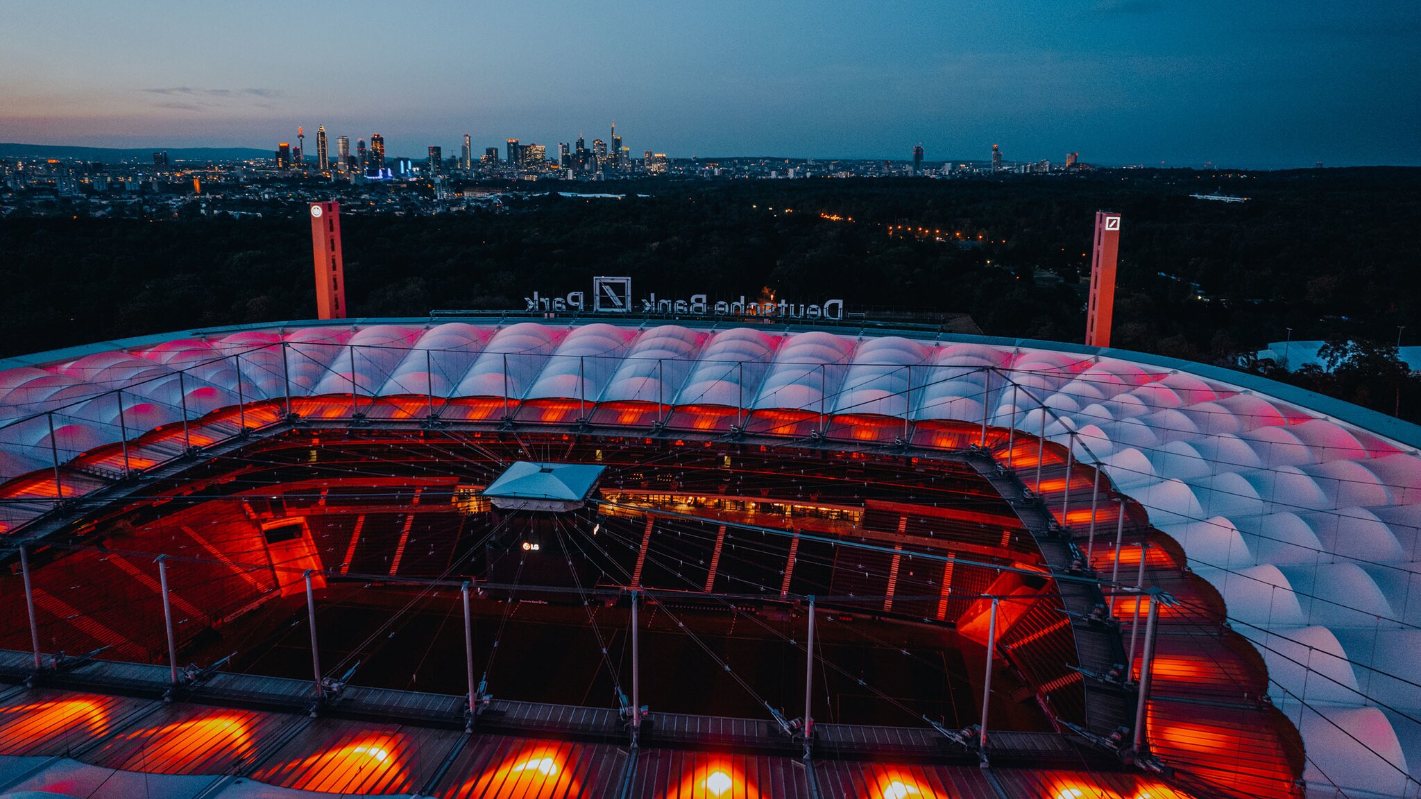 Luftaufnahme durch das geöffnete Dach des rot beleuchteten Deutsche Bank Park Stadions auf das Fußballfeld. Im Hintergrund die nächtliche Skyline von Frankfurt.