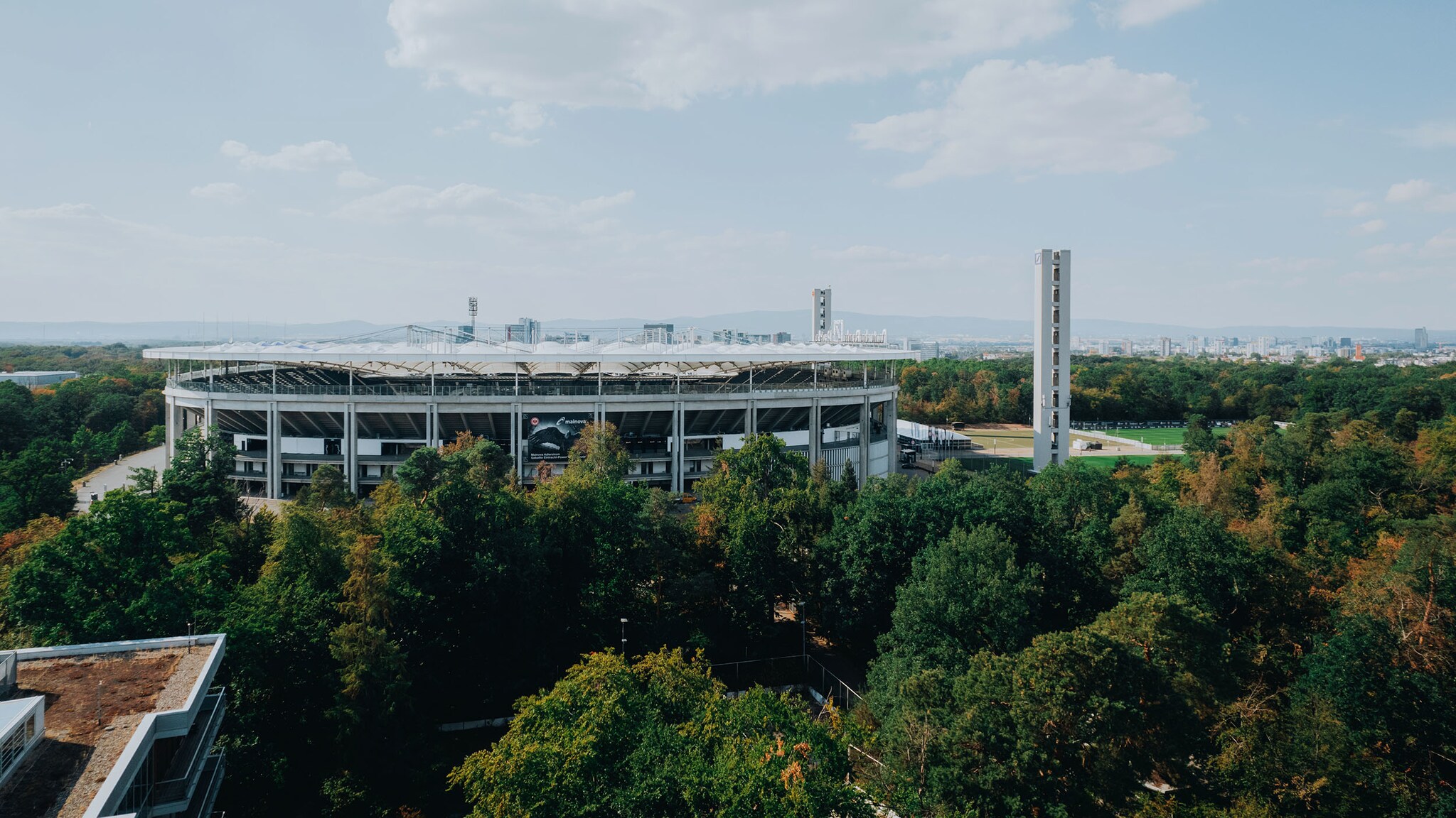 Luftaufnahme mit seitlichem Blick auf das Deutsche Bank Park Stadions in Frankfurt inmitten des Stadtwaldes, im Hintergrund die Skyline der Stadt.“