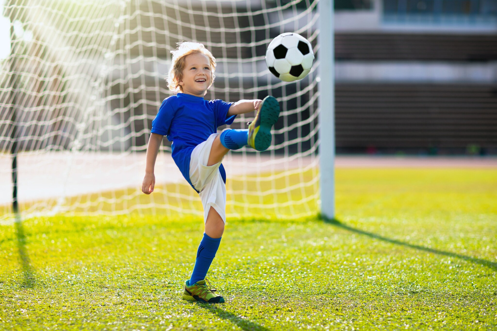 Ein lachender Junge in Sportkleidung kickt einen Fußball bei Sonnenschein hoch vor ein Tor auf dem Rasenplatz.