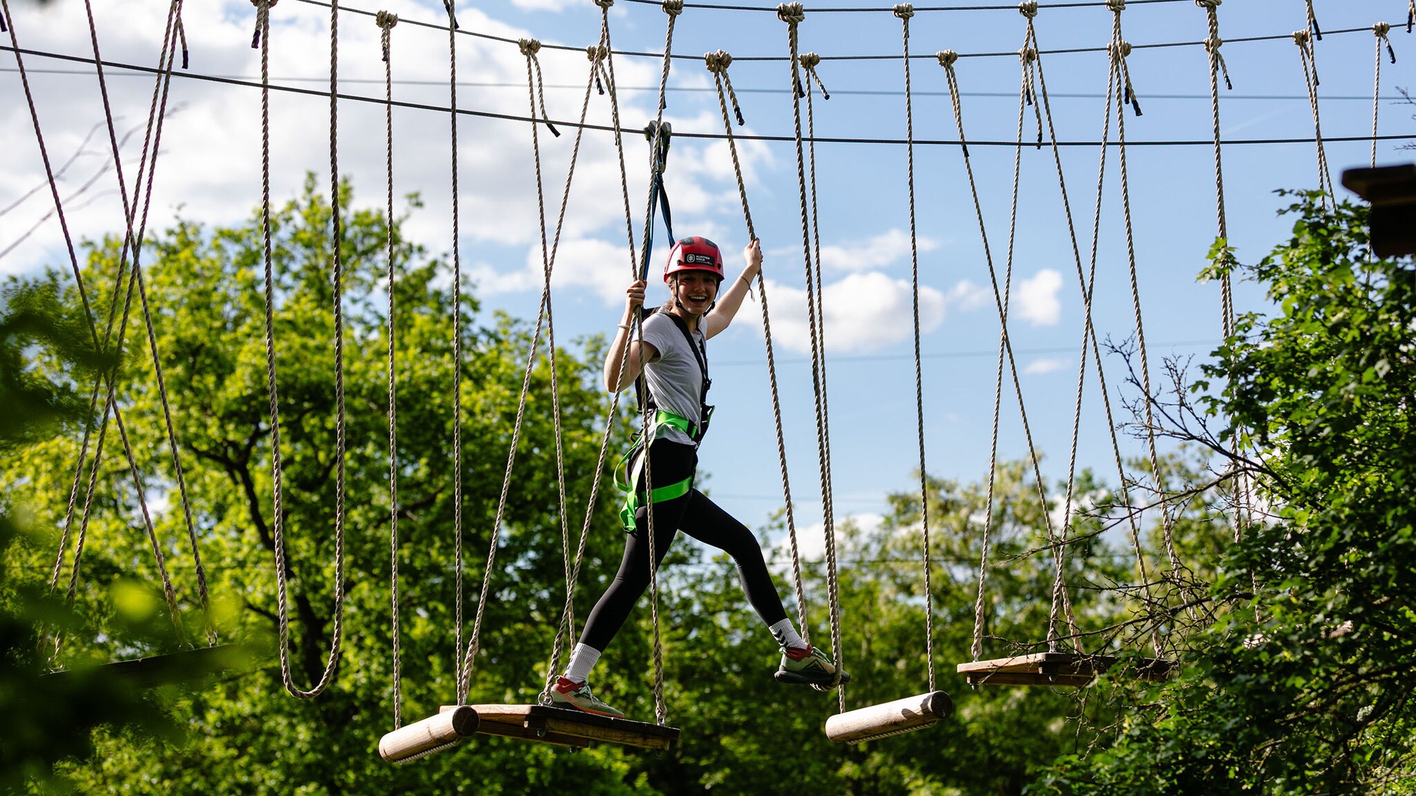 Ein Mädchen mit Helm läuft über eine Hängebrücke zwischen Baumwipfeln und schaut lachend in Richtung Kamera.