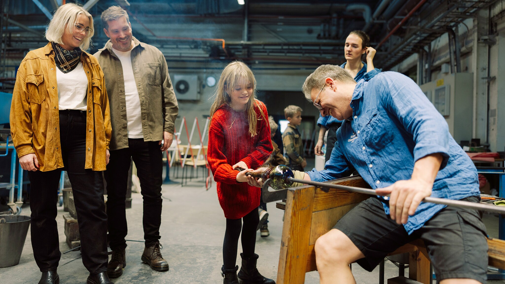 Eine Familie steht in einer Fabrikhalle und schaut einem Glasbläser bei der Arbeit zu, ein Mädchen hält dabei ein Ende einer Glasmacherpfeife fest. 