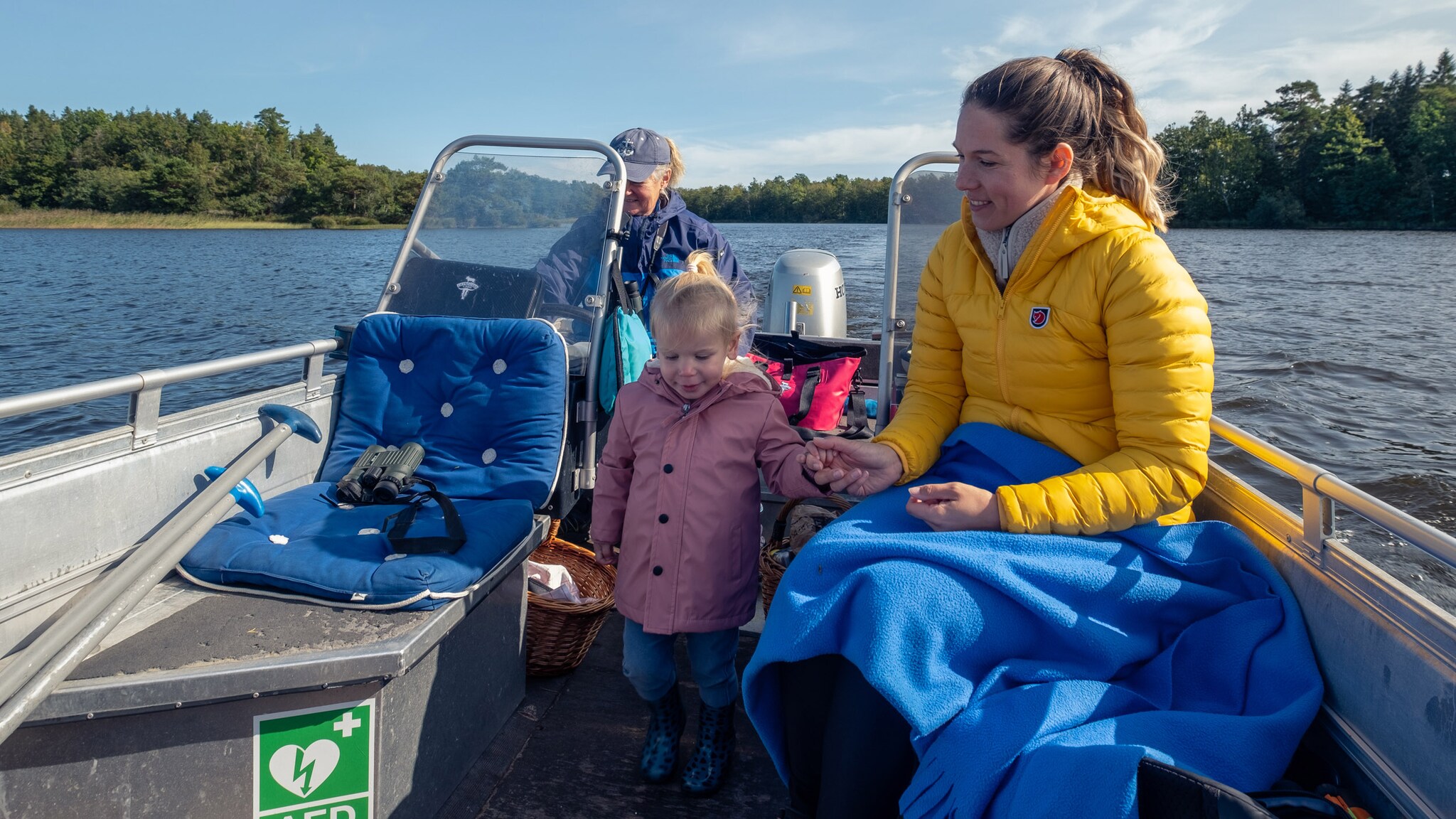 Zwei Frauen und ein Kind in Windjacken auf einem Motorboot auf einem See bei Sonnenschein.