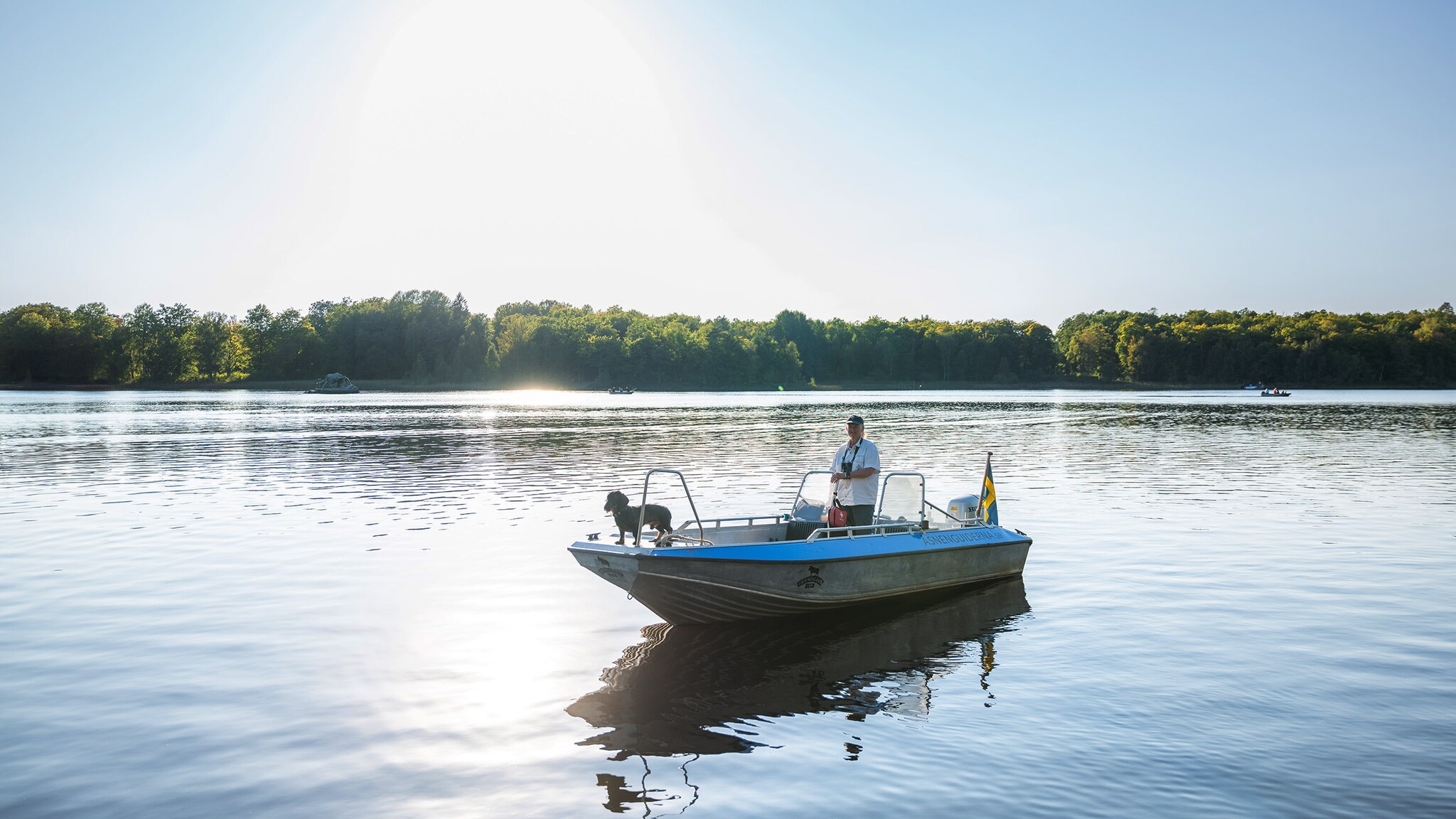 Eine Person und ein Hund stehen auf einem Motorboot auf einem See im Sonnenschein.