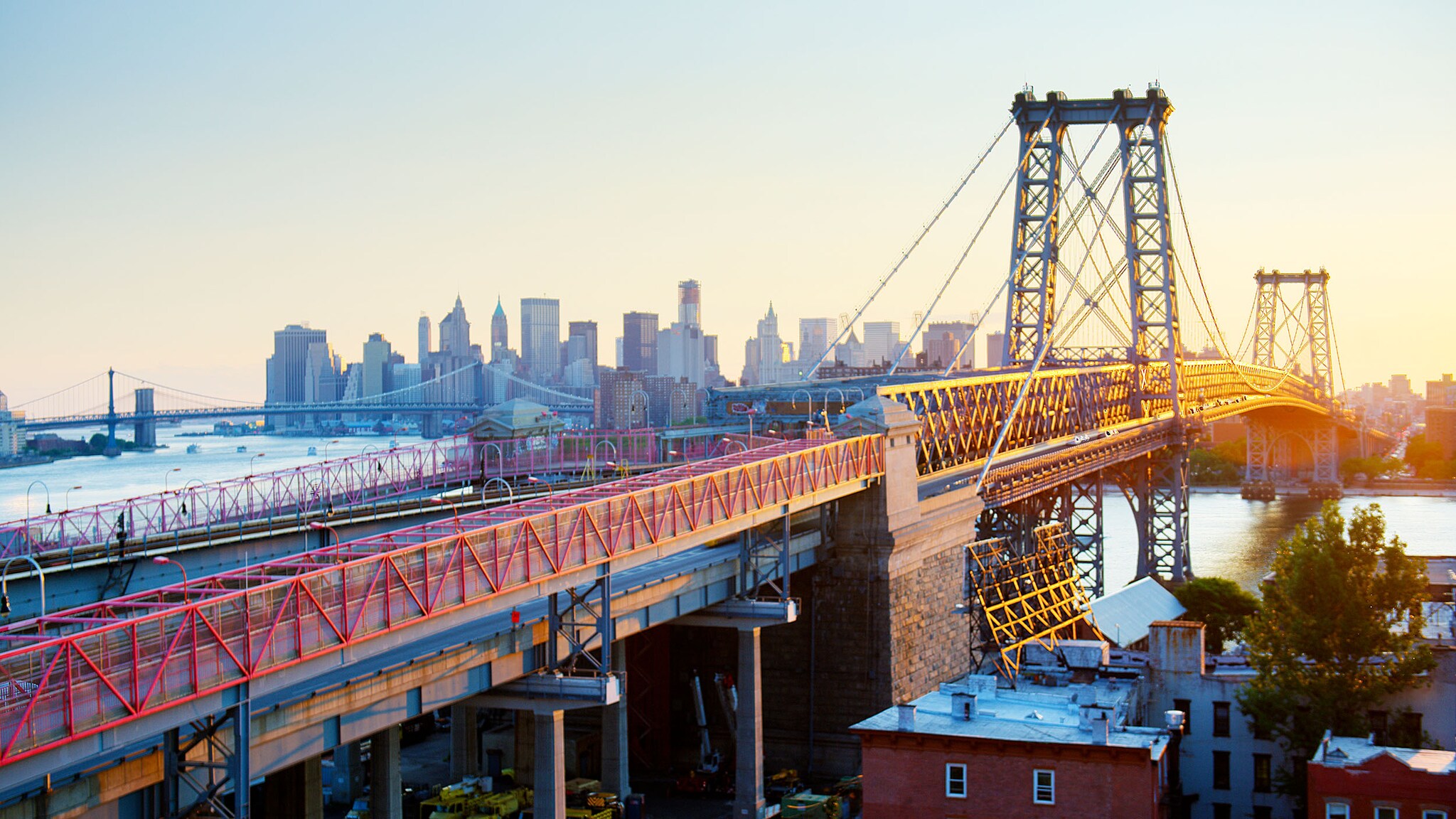 Stählerne Hängebrücke über Dächern und Fluss vor der Skyline von Manhattan bei Sonnenuntergang.