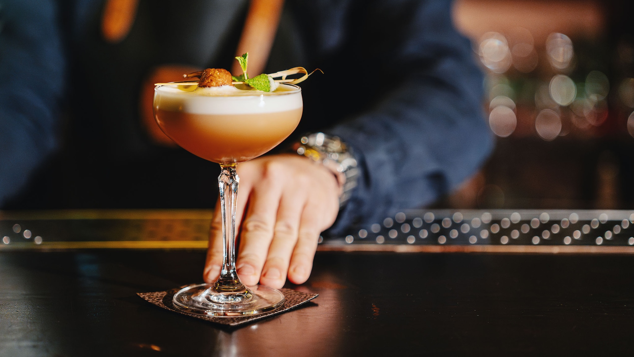 Eine Hand an einem garnierten Daiquiri-Cocktailglas auf einer Theke vor einer Person vor unscharfen Hintergrund.