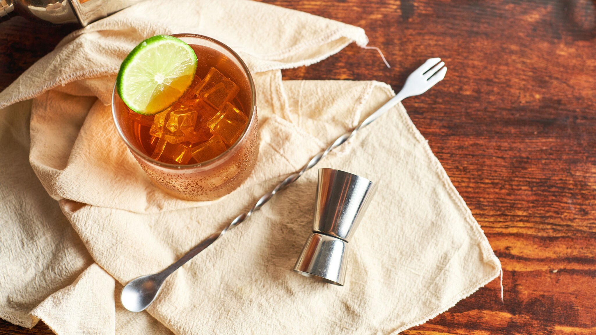 Aufsicht auf einen hellbraunen Longdrink mit Eiswürfeln, garniert mit einer Limettenscheibe, auf einem Tuch neben Bar-Accessoires.