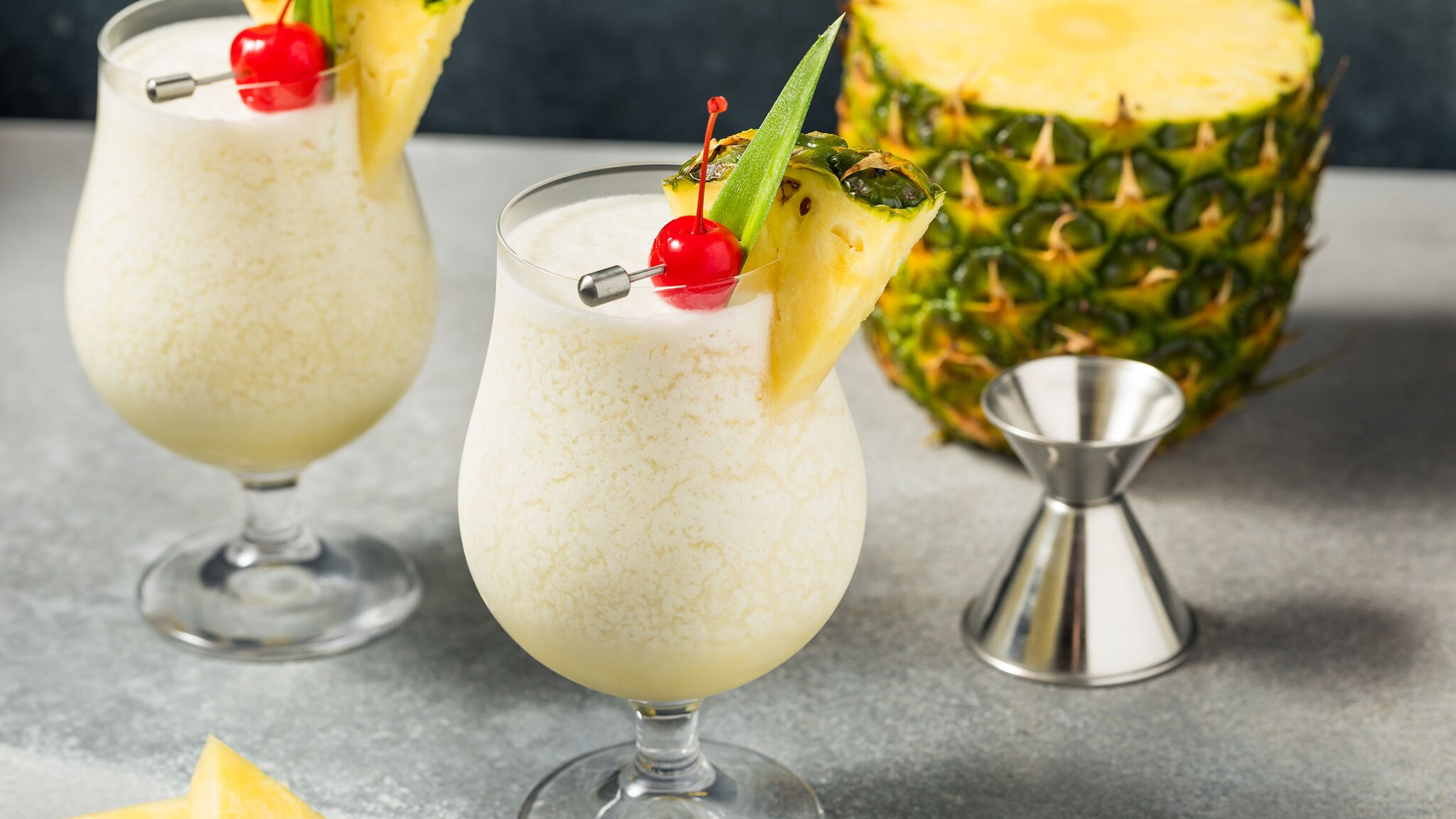 Zwei cremige, helle Pina-Colada-Cocktails in bauchigen Cocktailgläsern, garniert mit Ananasspalte und Cocktailkirsche, auf einem silberfarbenen Untergrund, dahinter eine Ananas.