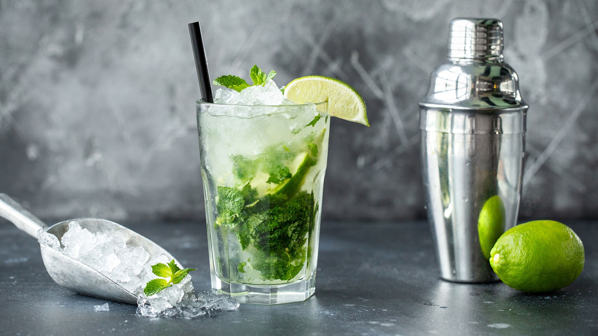 Ein Glas mit Mojito auf Eis mit Minze und Limette, angerichtet auf einem silberfarbenen Untergrund neben einer Eisschaufel und einem Cocktailshaker.