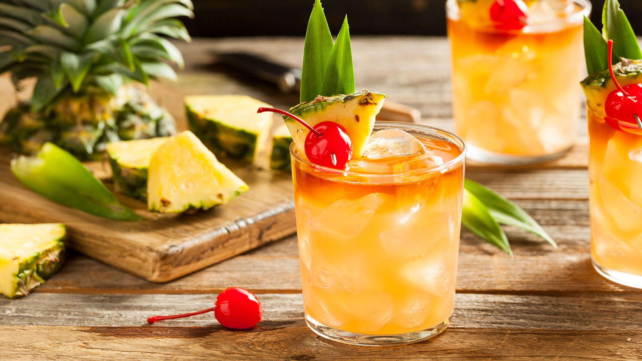 Drei Gläser mit orangefarbenen Mai-Tai-Cocktails mit Eiswürfeln, garniert mit Ananasspalte und Cocktailkirsche auf einem Holztisch, dahinter Ananasstücke.