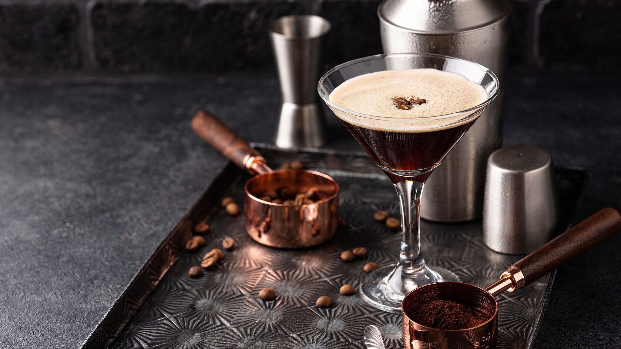 Ein Espresso-Cocktail mit weißer Crema und Kaffeebohnen in einem Martiniglas, arrangiert auf einem silbernen Tablett mit Cocktailshaker und Kaffee-Accessoires.