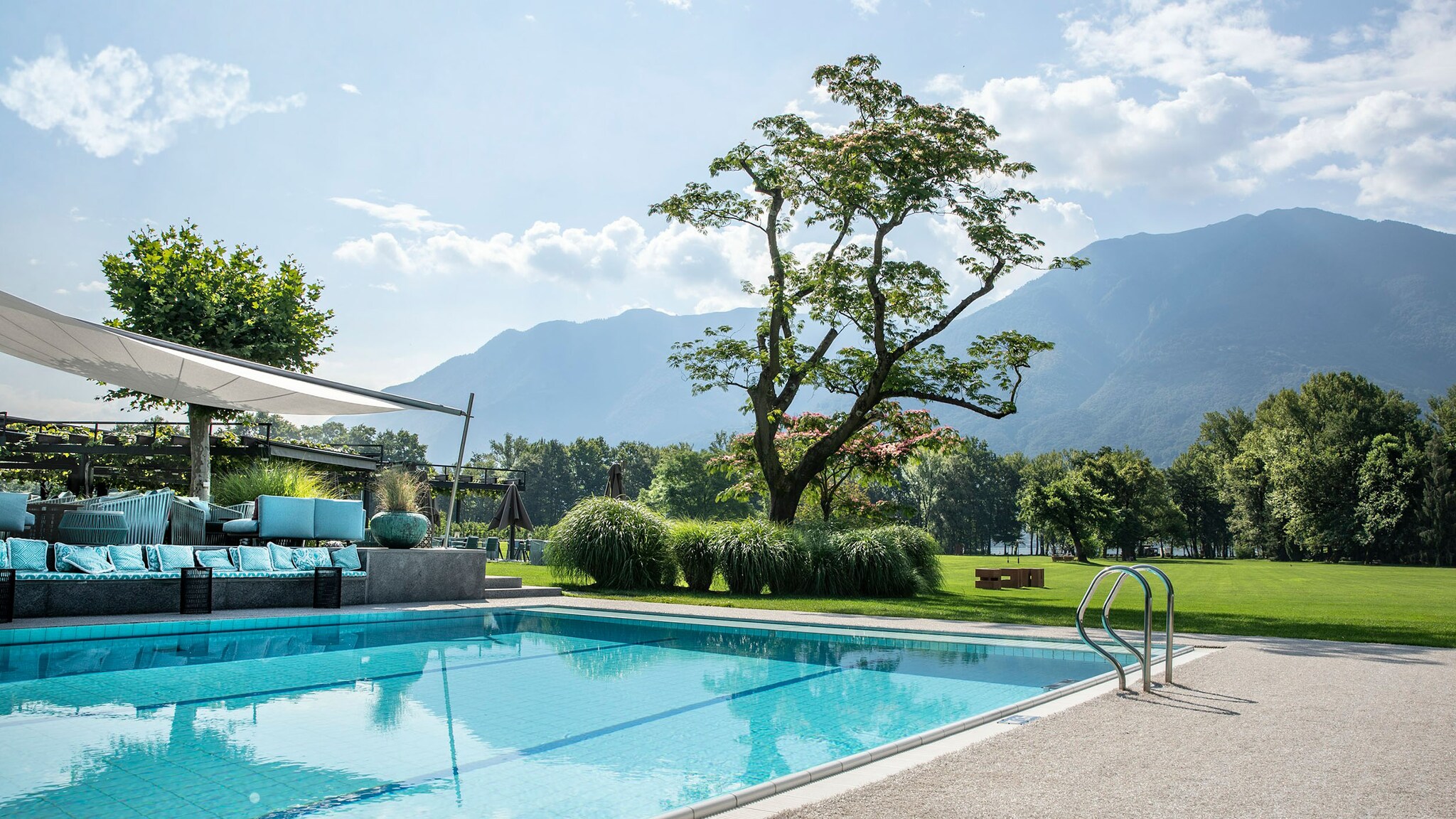 Terrasse mit Sonnenliegen und Pool in einer Gartenanlage mit Rasenfläche vor Bergpanorama.