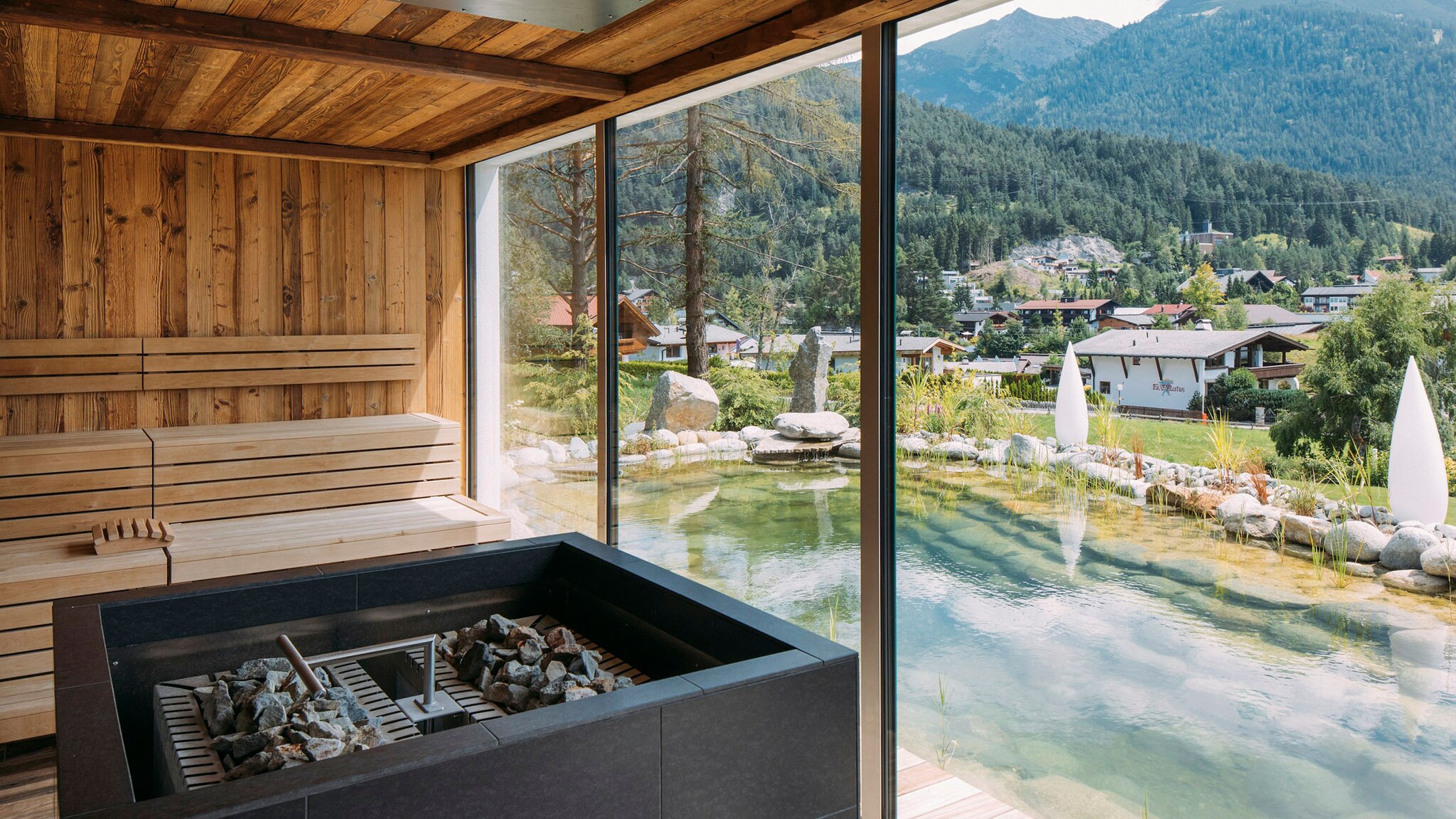Blick aus einer Holzsauna mit Ofen über einen Schwimmteich in eine Berglandschaft.