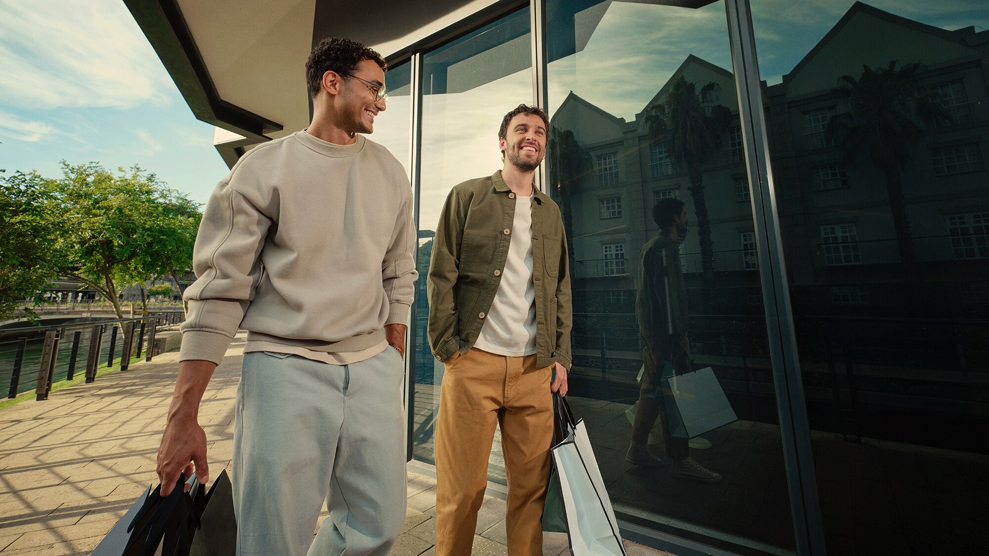 Zwei lächelnde Männer laufen lässig mit Einkaufstüten in den Händen vor einer Gebäudefront aus Glas in urbanem Setting.