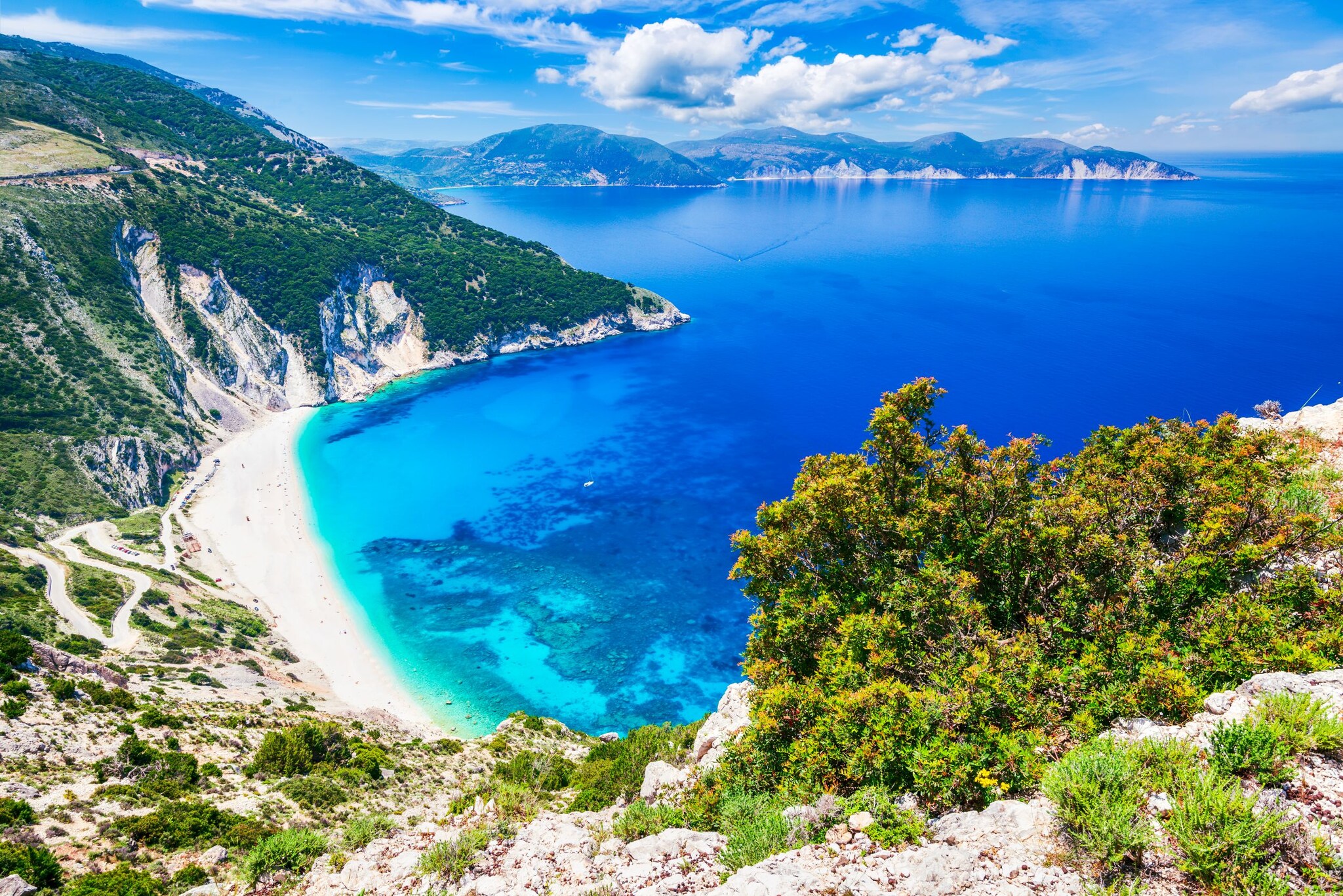 Blick von oben auf einen weißen Sandstrand und blaues Meer an einer felsigen Küste. 