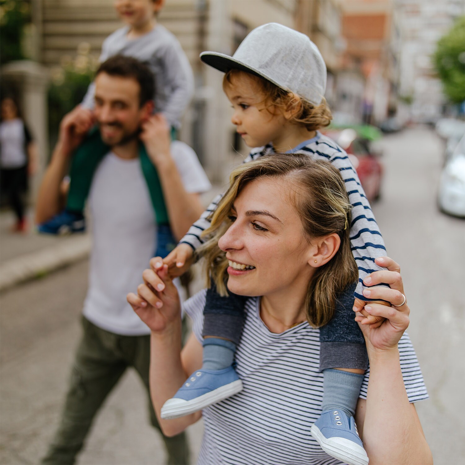 Eine Frau und ein Mann gehen gut gelaunt mit Kindern auf ihren Schultern auf einer Straße in einer Stadt.
