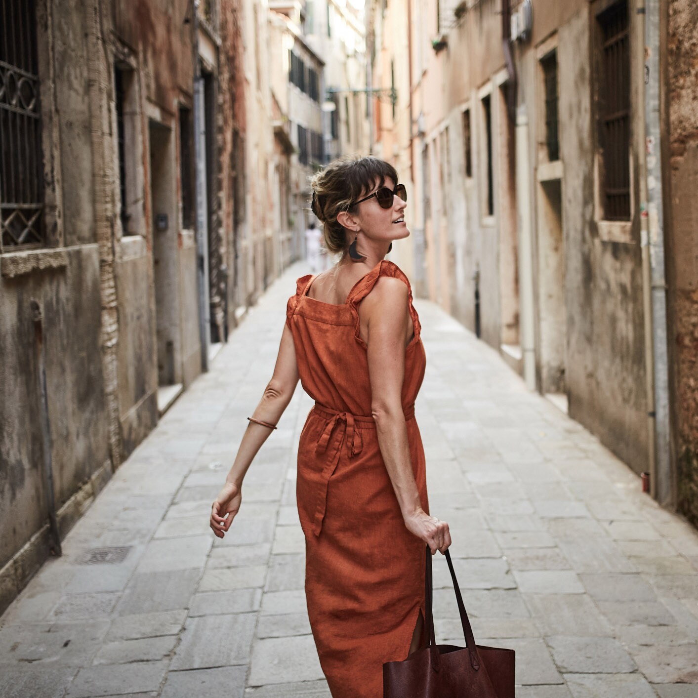 Rückansicht einer Frau im Sommerkleid und mit Sonnenbrille, die gut gelaunt mit einem Trolley durch eine Gasse in einer mediterranen Altstadt läuft.