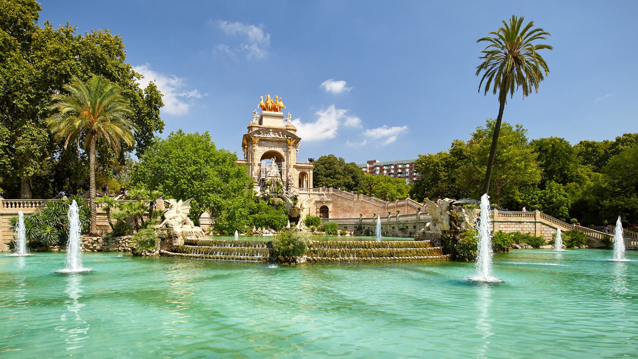 Parkanlage mit monumentalen Brunnen mit goldener Quadriga bei Sonnenschein.