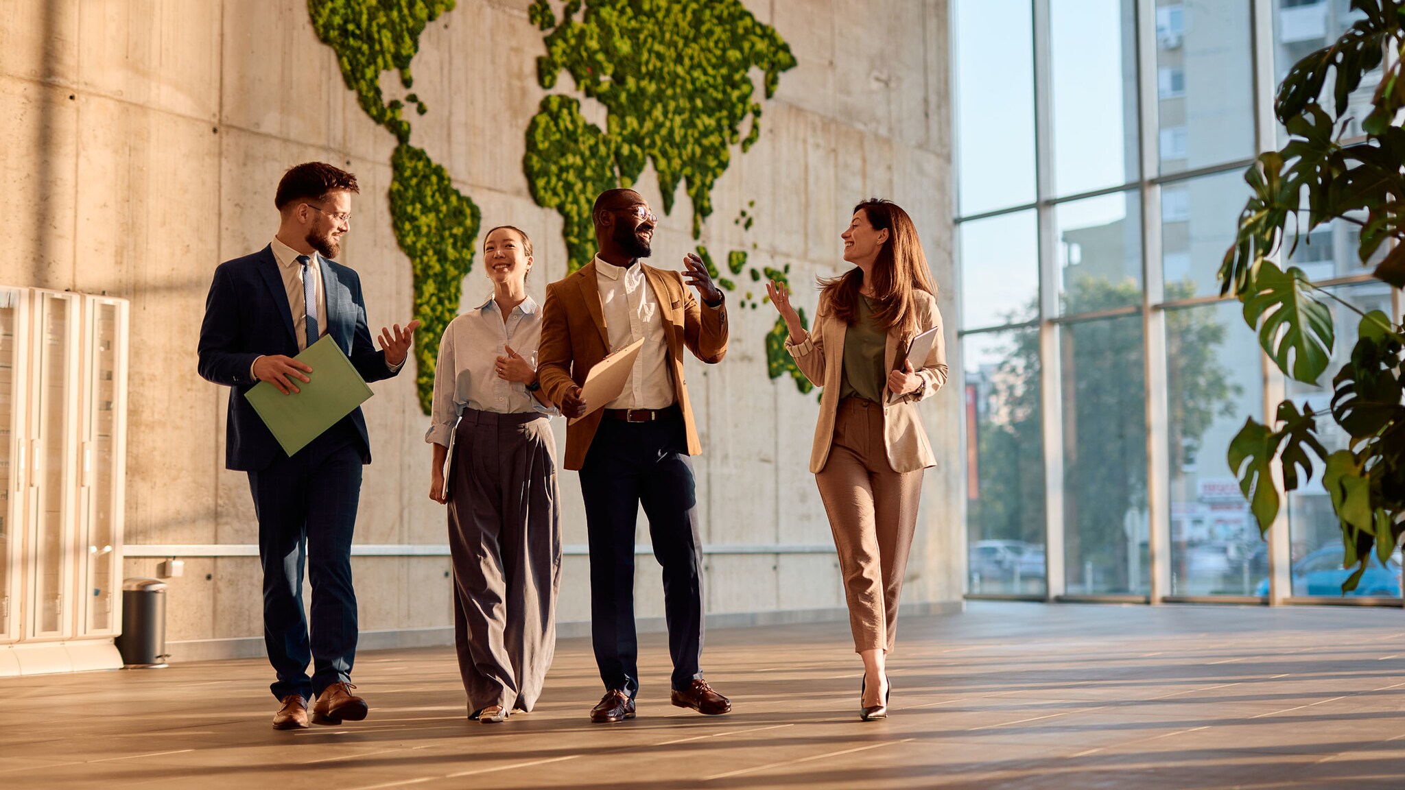 Zwei Frauen und zwei Männer in Business-Kleidung gehen gut gelaunt mit Arbeitsmappen durch ein Foyer vor einer Betonwand mit grüner Weltkarte.