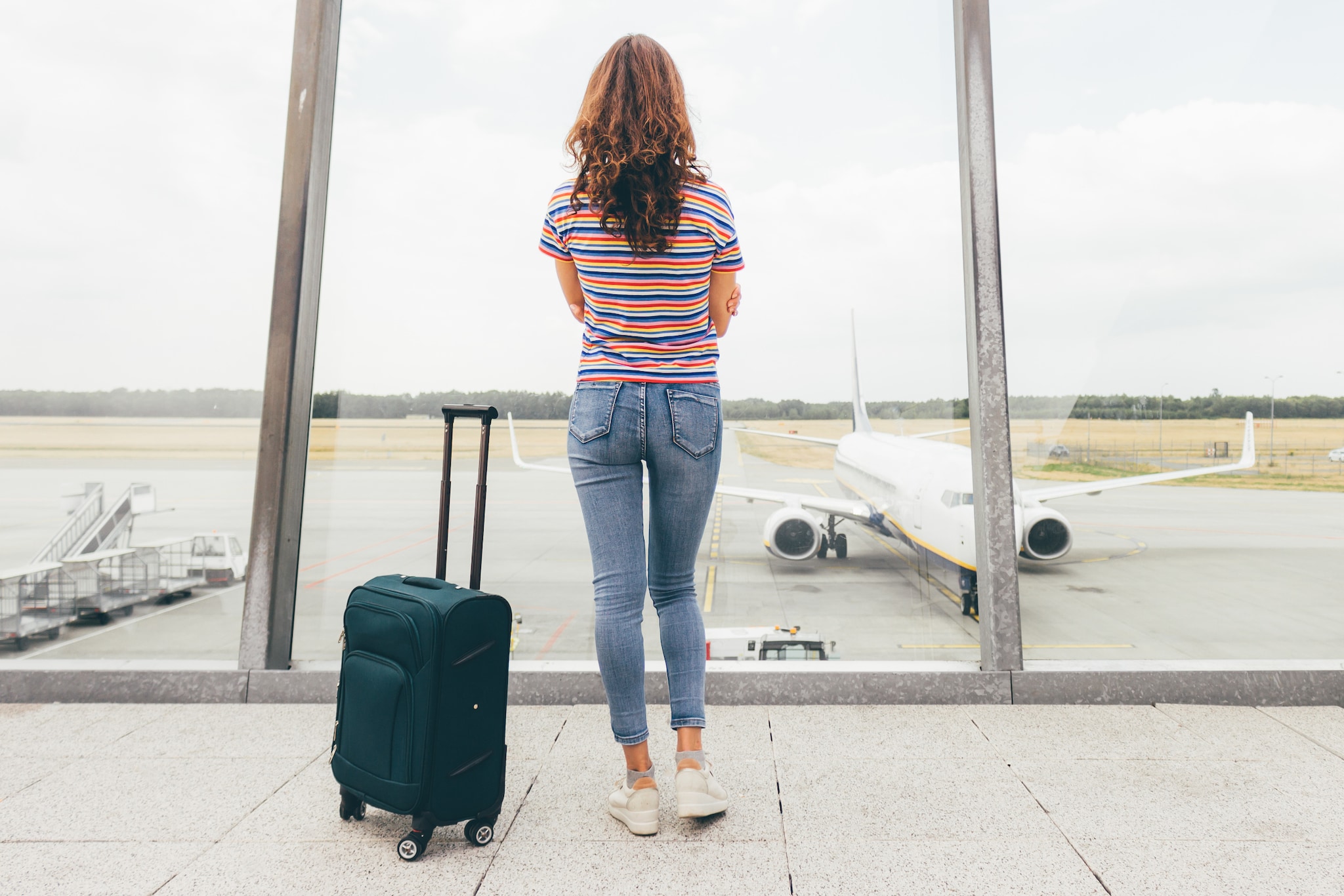 amex-insurance-flug-annulliert-ersatzflug-frau-koffer Rückansicht einer Frau mit langen braunen Haaren, in Jeans und gestreiftem T-Shirt vor einer Glasscheibe an einem Flughafen mit einem Flugzeug, neben ihr ein Rollkoffer