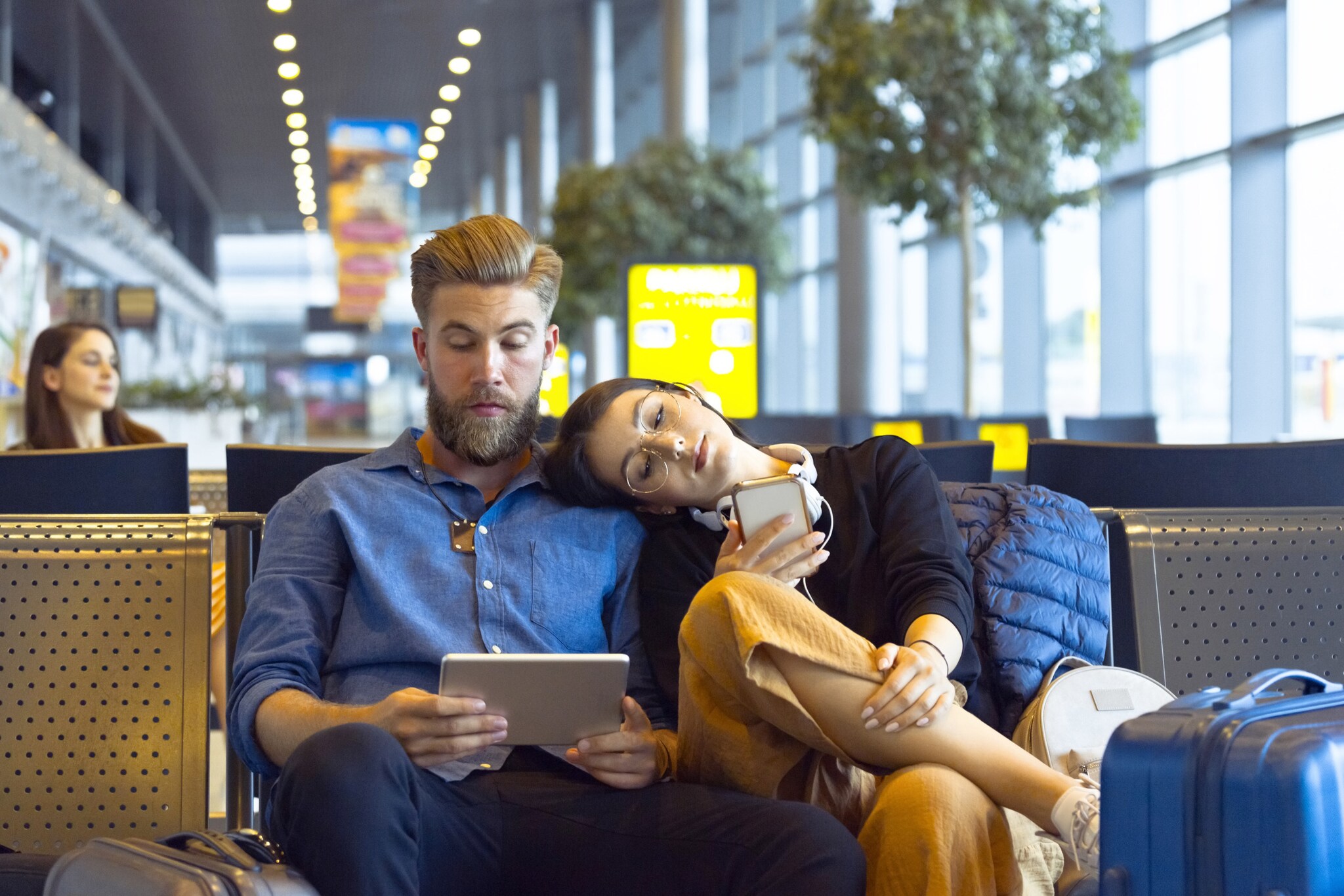 Ein junger Mann und eine junge Frau sitzen im Wartebereich eines Flughafens, ihr Kopf ruht auf seiner Schulter, beide schauen auf ihre mobilen Endgeräte