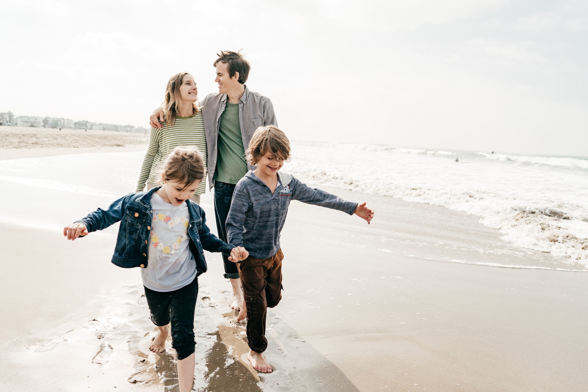 amex-insurance-reiseversicherung-wichtigste-info-familie Eine Familie läuft am Strand, die beiden Kinder Hand in Hand vor ihren Eltern, die sich lächelnd anschauen.