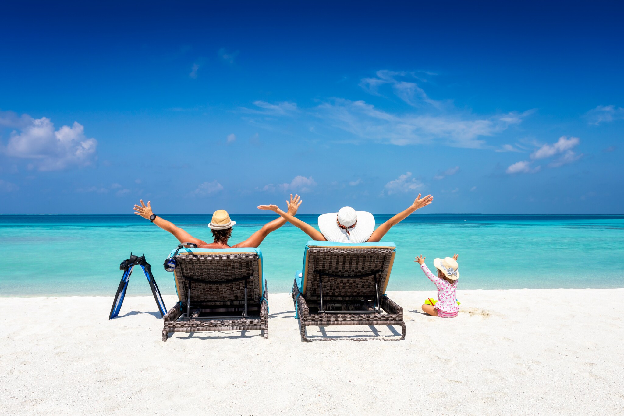 Eine Familie, die von hinten zu sehen ist, liegt auf Sonnenliegen am Strand und hält die Arme in die Luft.
