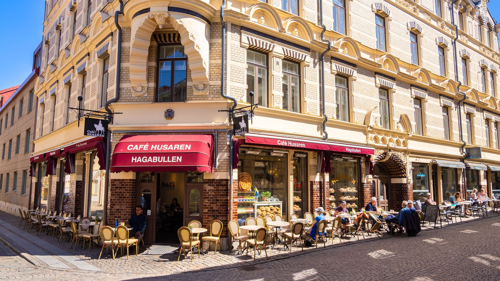 Personen in einem Straßencafé mit roter Markiese in einem historischen Eckhaus bei Sonnenschein.