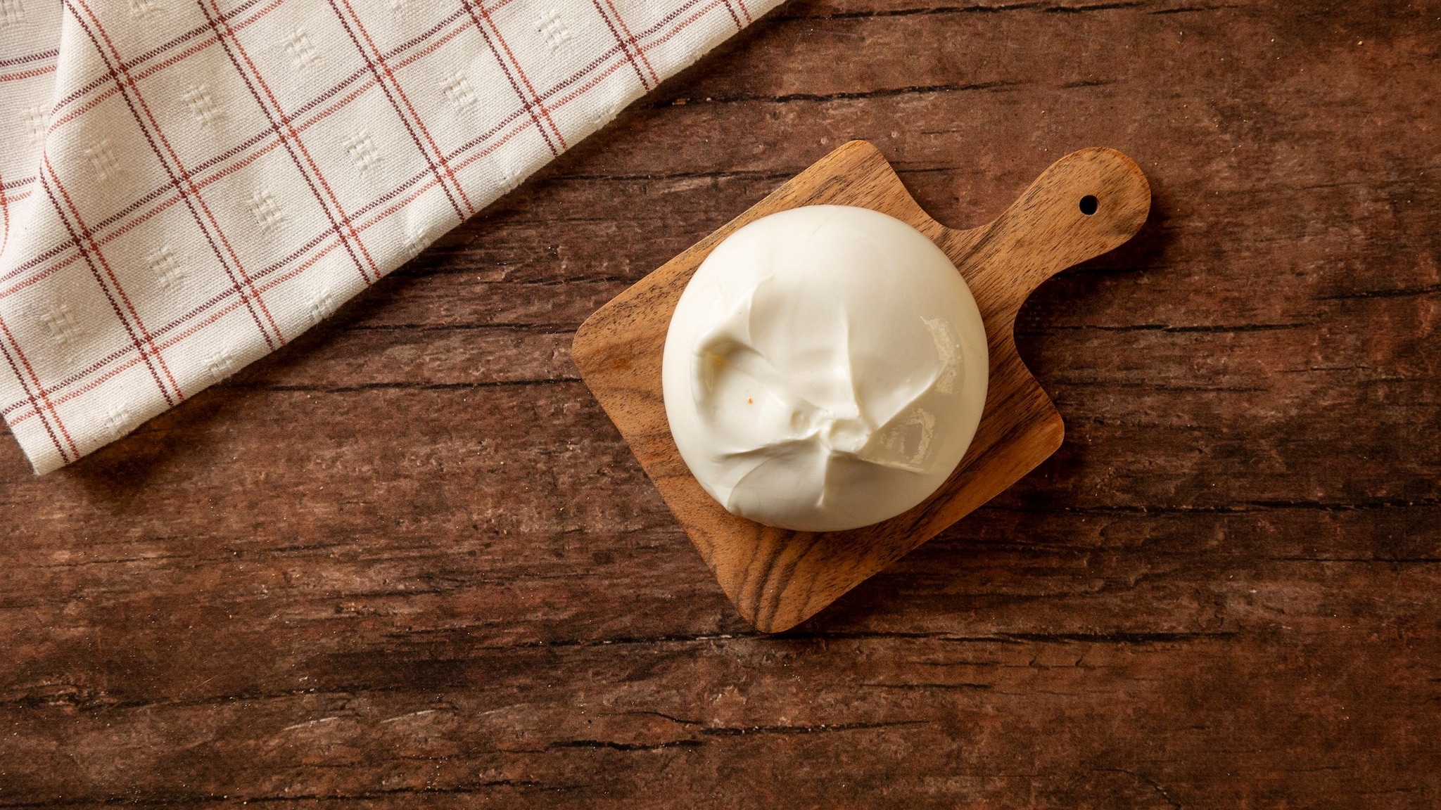 Ein runder Burrata auf einem Holzbrett, das mit einem karierten Küchentuch auf einem Holztisch liegt.