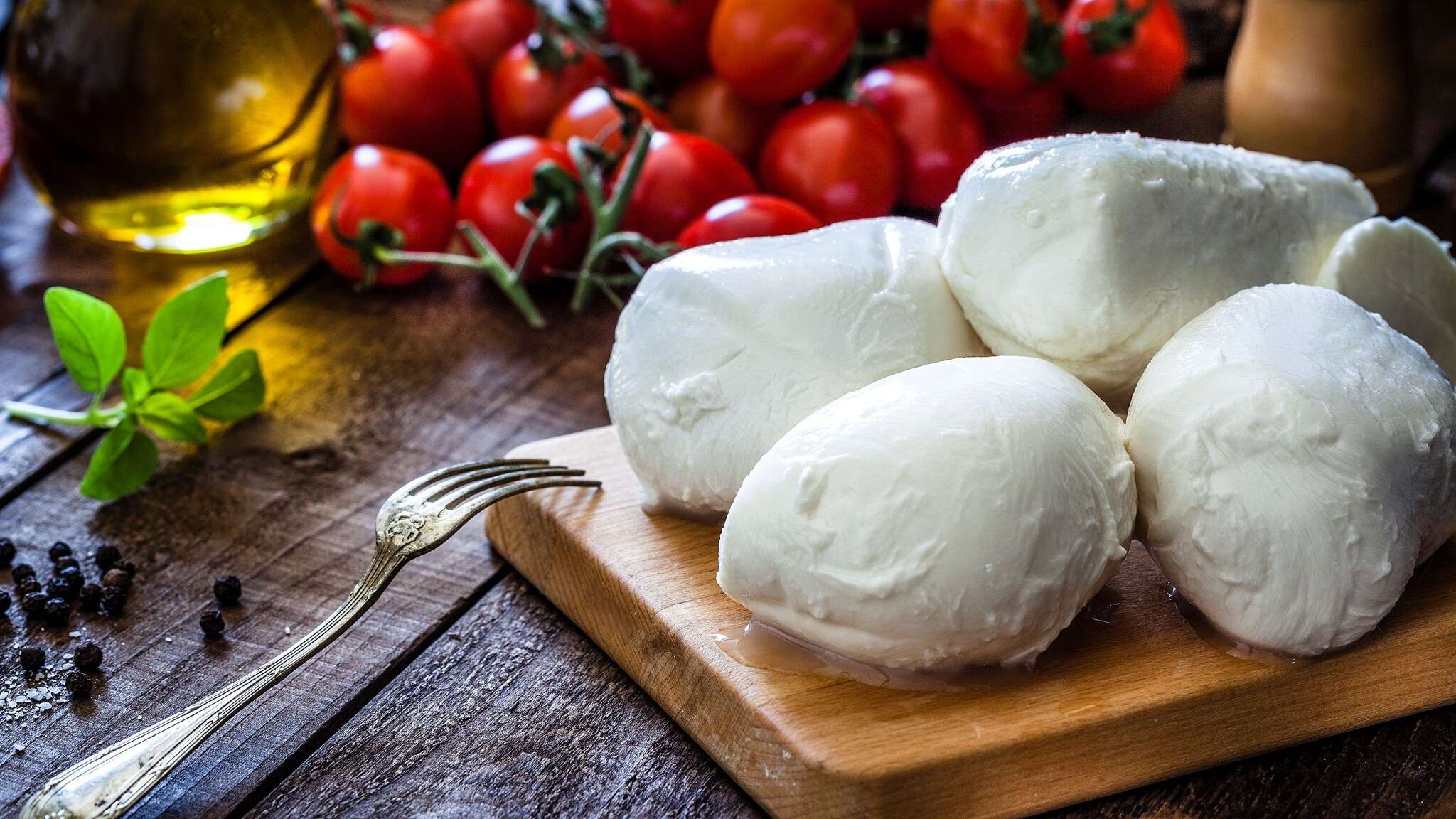 Runder Mozzarella-Käse auf einem Holzbrett, das mit Tomaten, Gewürzen und einer Gabel auf einem Holztisch liegt.