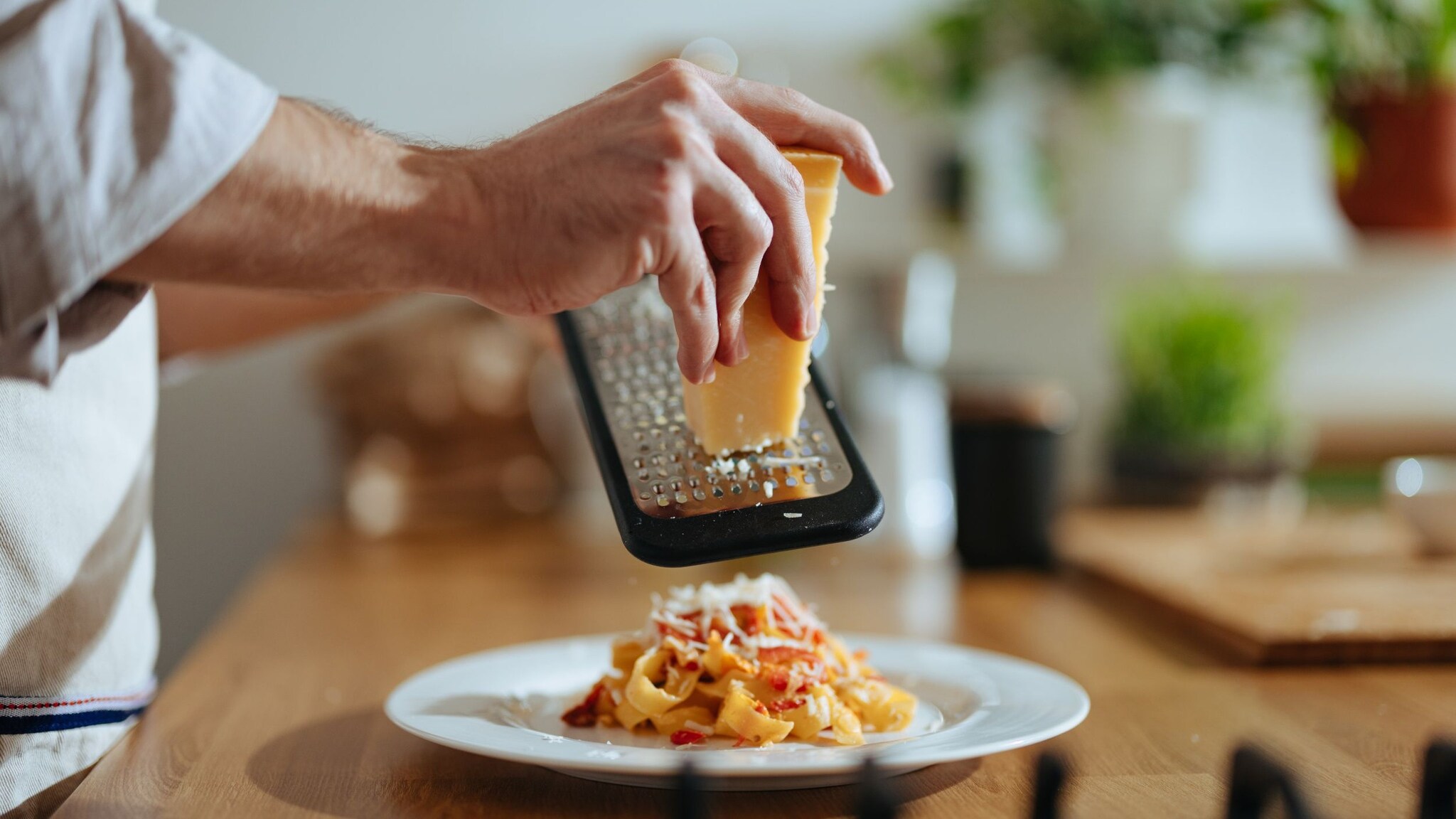 Nahaufnahme von Händen, die mit einer Reibe Käse auf einen Teller mit Pasta reiben.