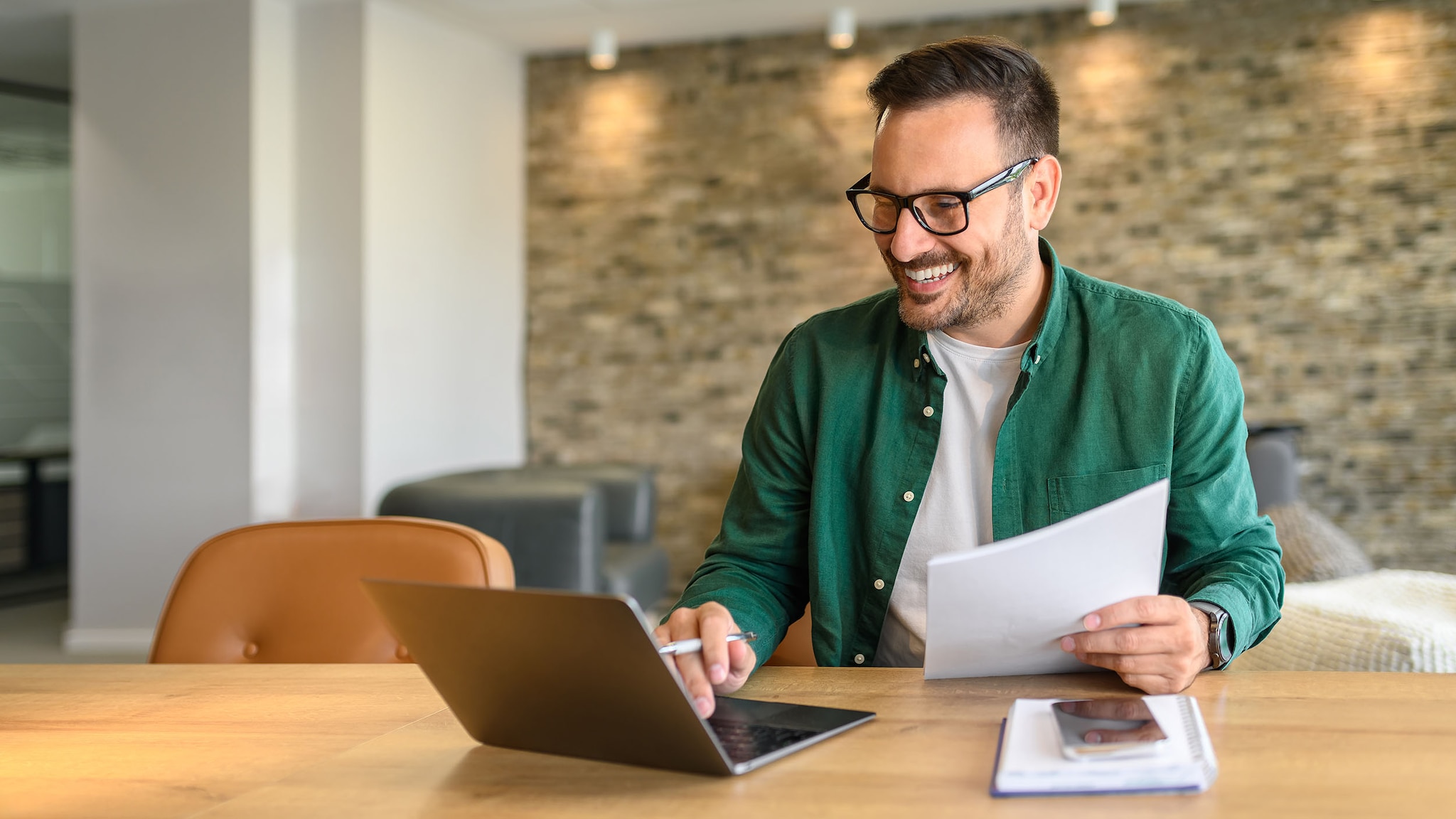 Ein Mann im grünen Hemd sitzt an einem Schreibtisch und arbeitet gut gelaunt mit Arbeitsunterlagen in der Hand an einem Laptop in einem Büro.