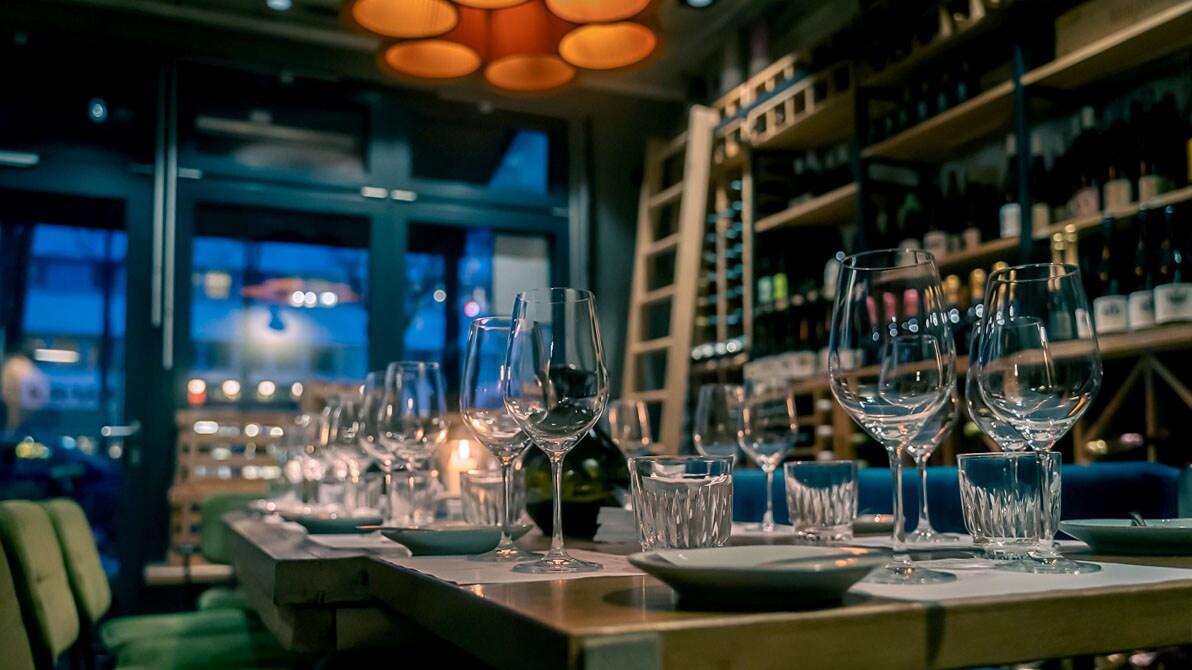 In einem Restaurant steht ein großer Tisch mit vielen Weingläsern, im Hintergrund sieht man ein Weinregal.
