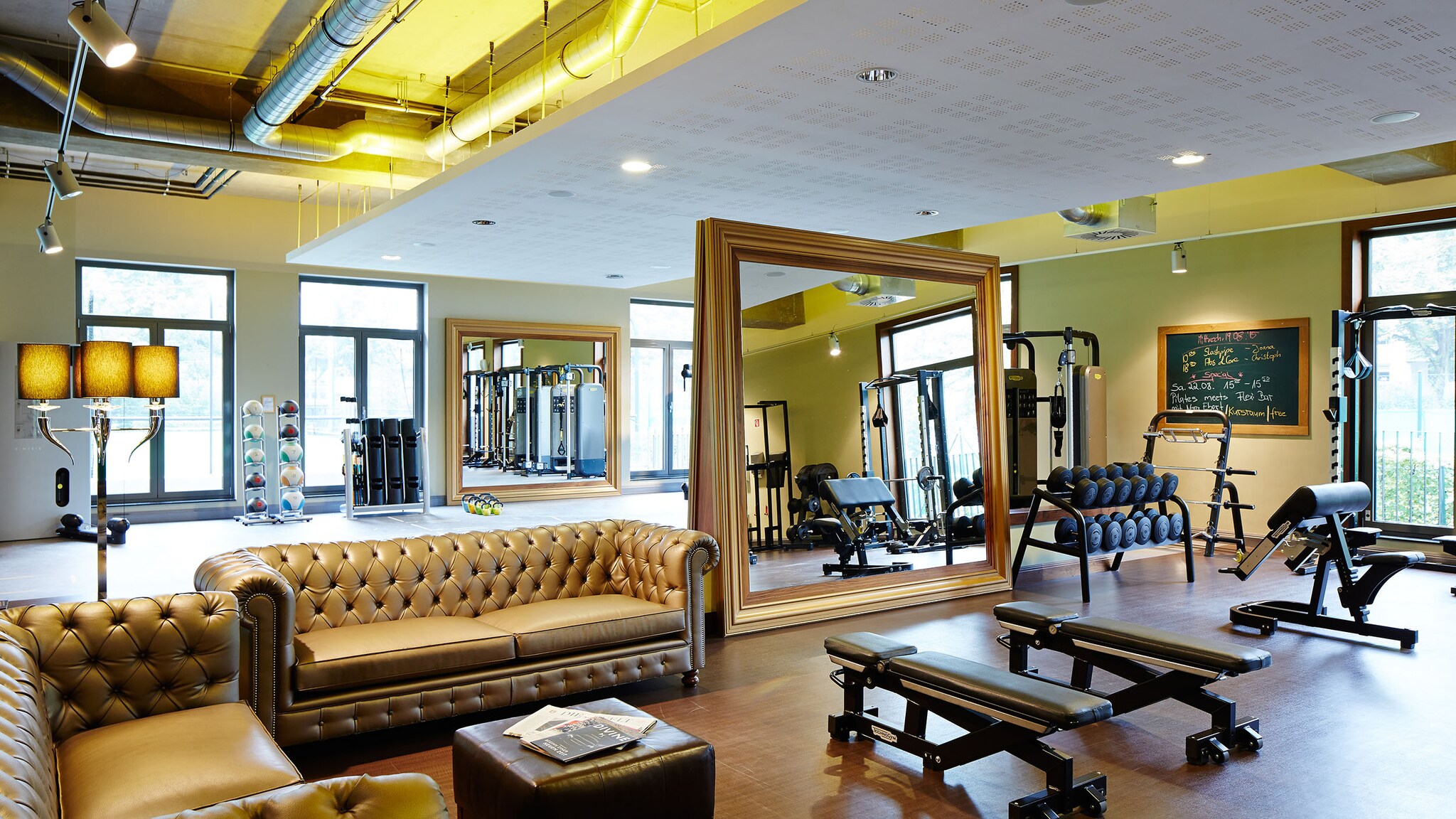 In einem Fitnessraum steht ein großer Spiegel mit Goldrahmen, umgeben von Sportgeräten und Sofas.