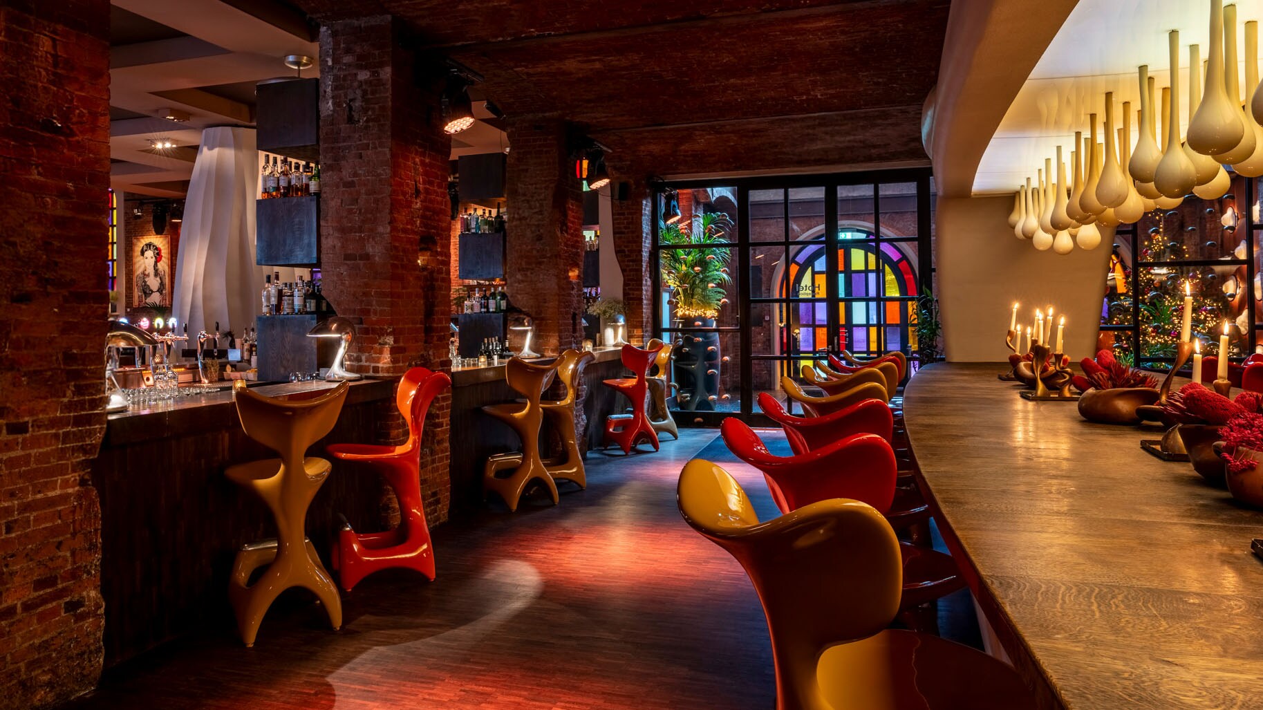 In einer Hotel-Lobby stehen bunte Stühle am einer Bar, im Hintergrund ist eine Tür aus buntem Glas.