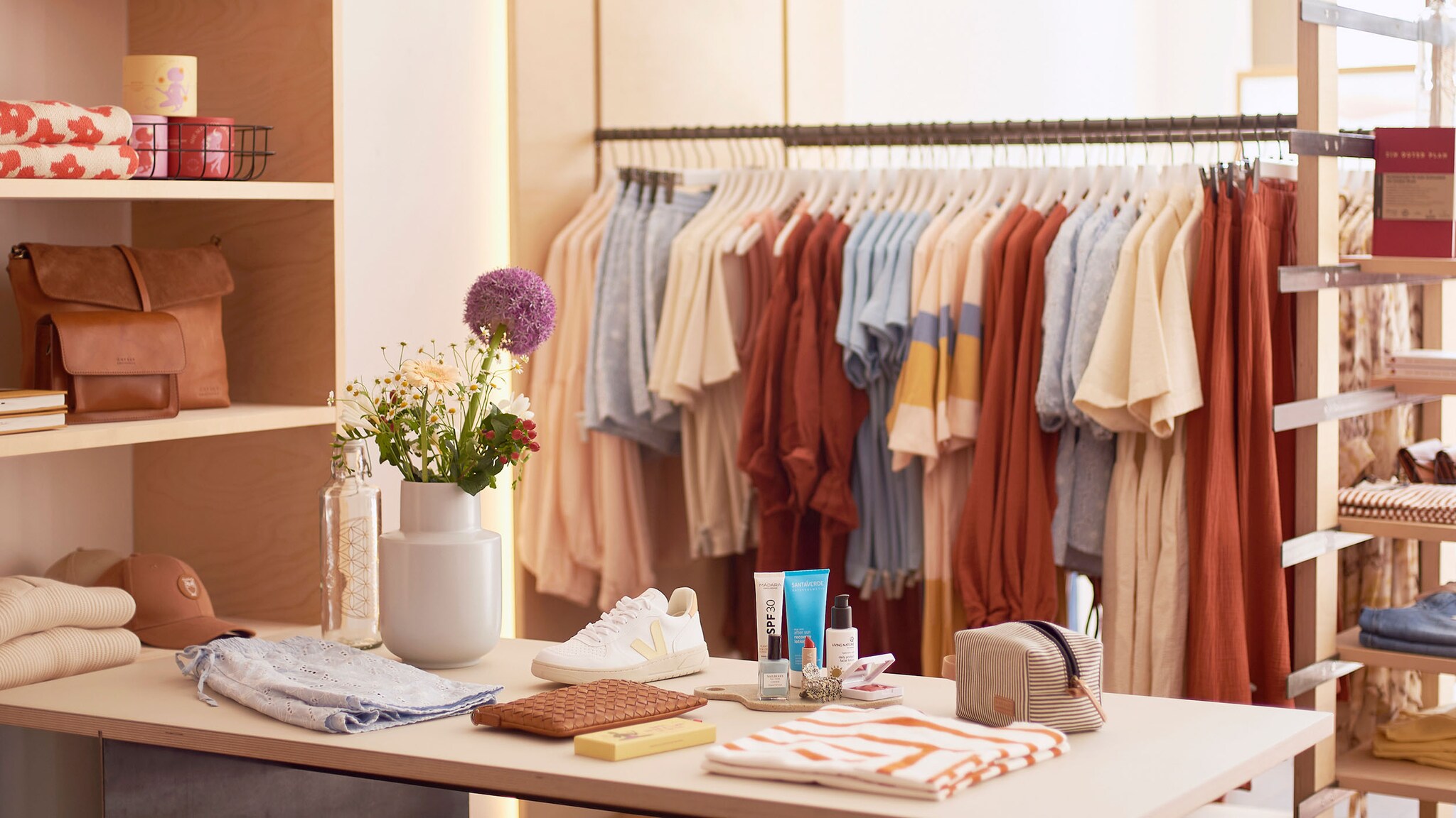 In einem Laden stehen Ständer mit Kleidung, davor ist ein Tisch mit Sneakern, Kosmetik und einer Blumenvase.