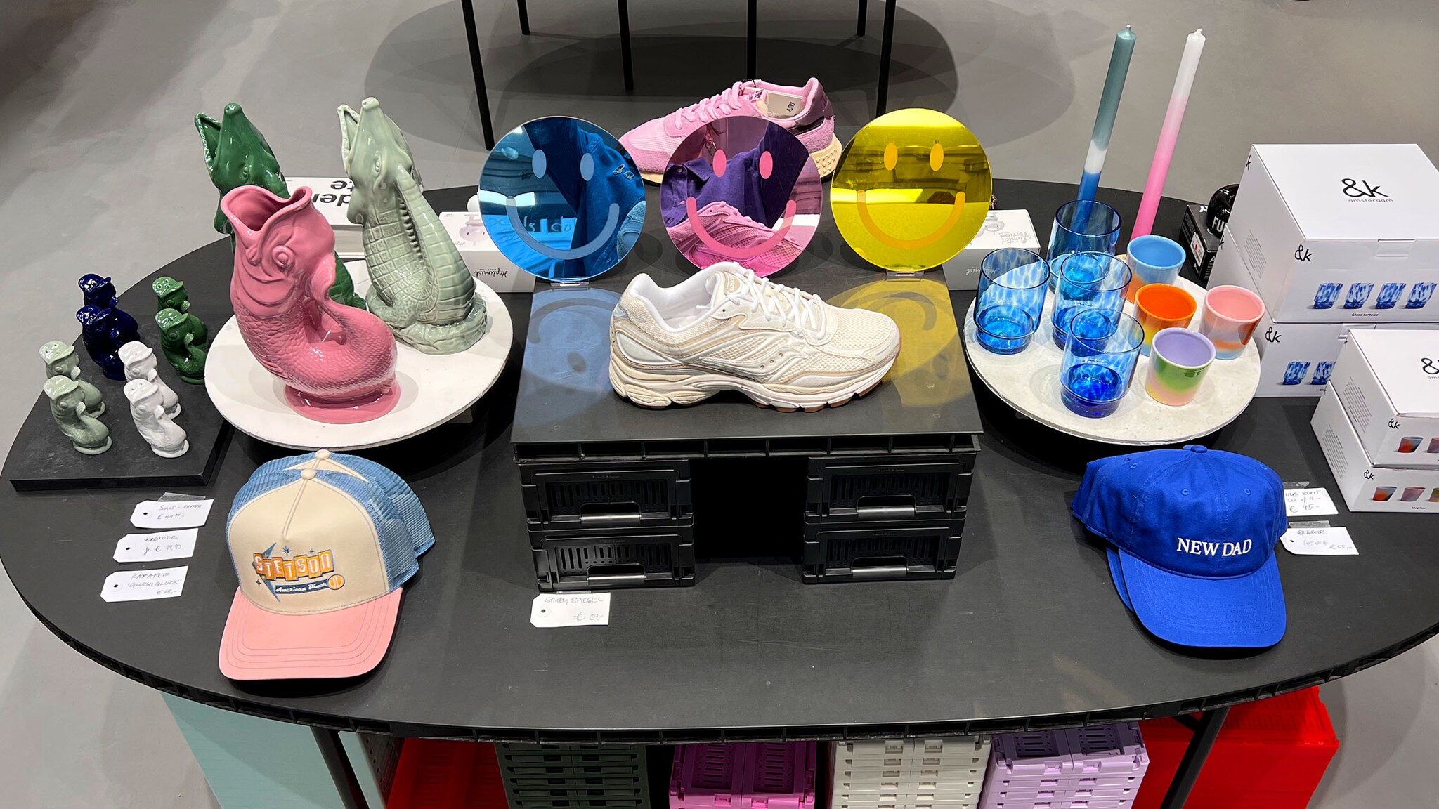 Auf einem schwarzen Tisch stehen bunte Fischvasen, Caps, Spiegel in Smiley-Form und ein weißer Sneaker. 