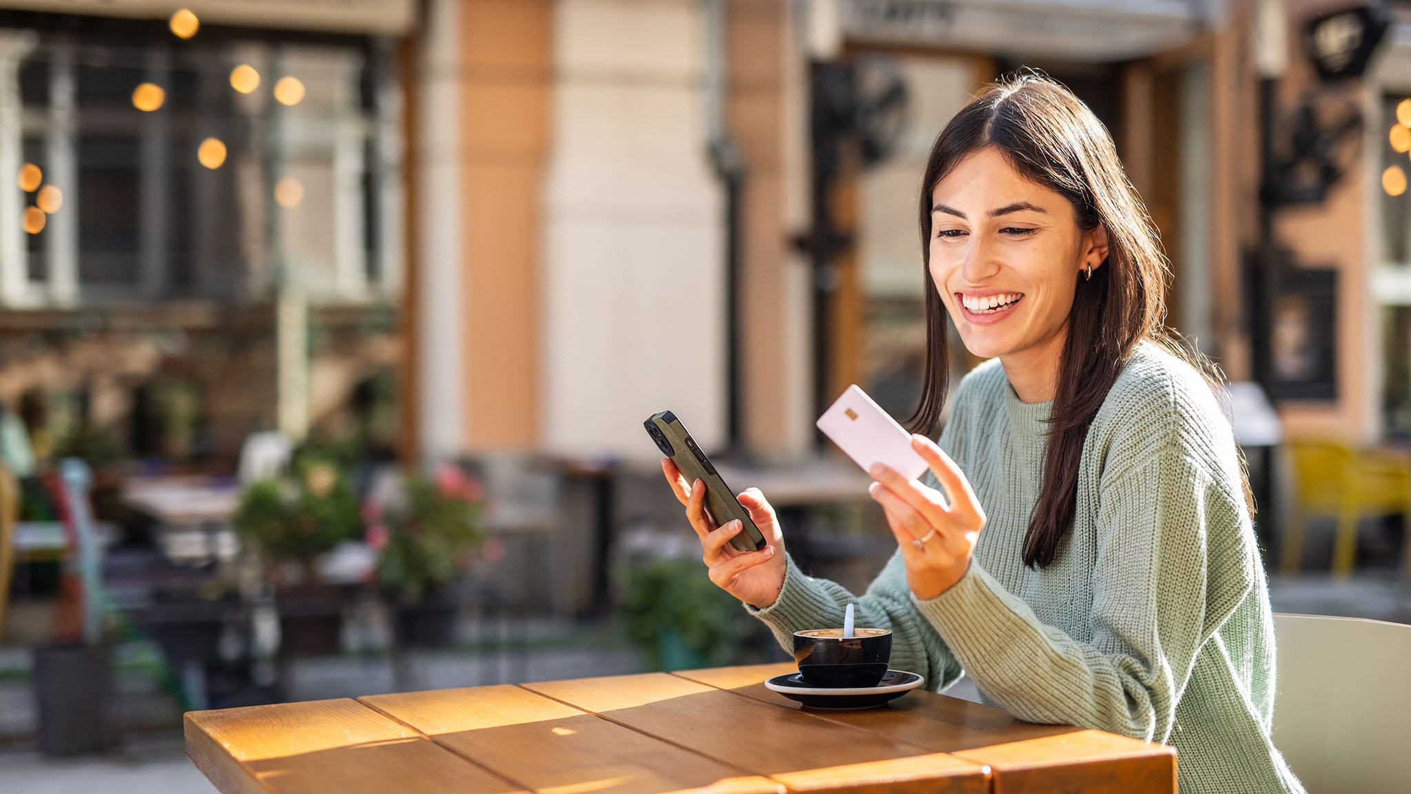 Eine Frau sitzt mit dem Smartphone in der Hand am Tisch und schaut auf ihre Kreditkarte.