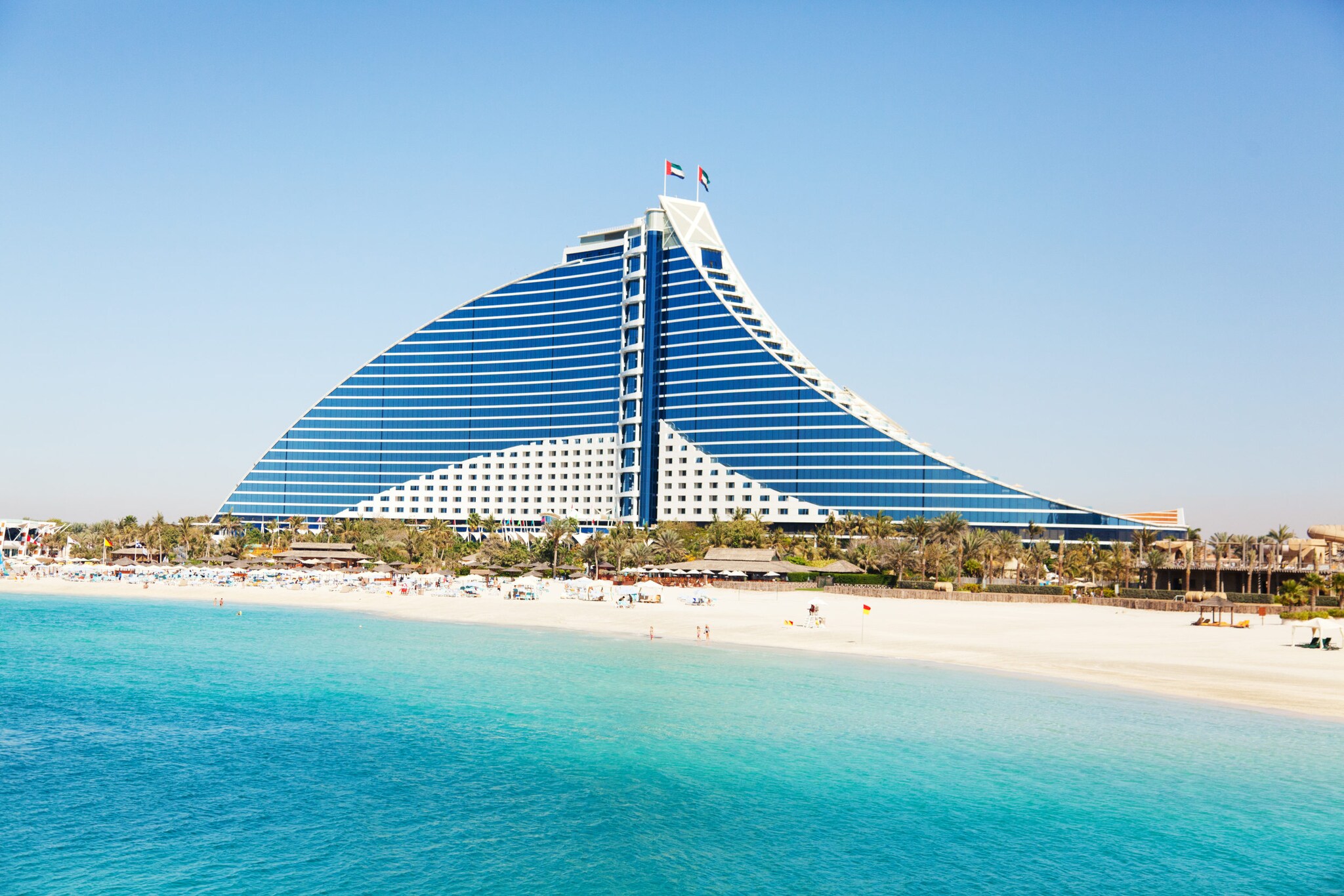 Blick auf das moderne wellenförmige Jumeirah Beach Hotel, mit Strand und blauem Wasser im Vordergrund.