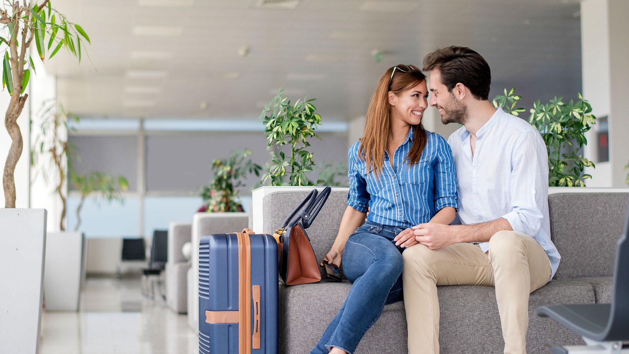 Eine Frau und ein Mann sitzen in einer Lounge am Flughafen und lächeln sich an.