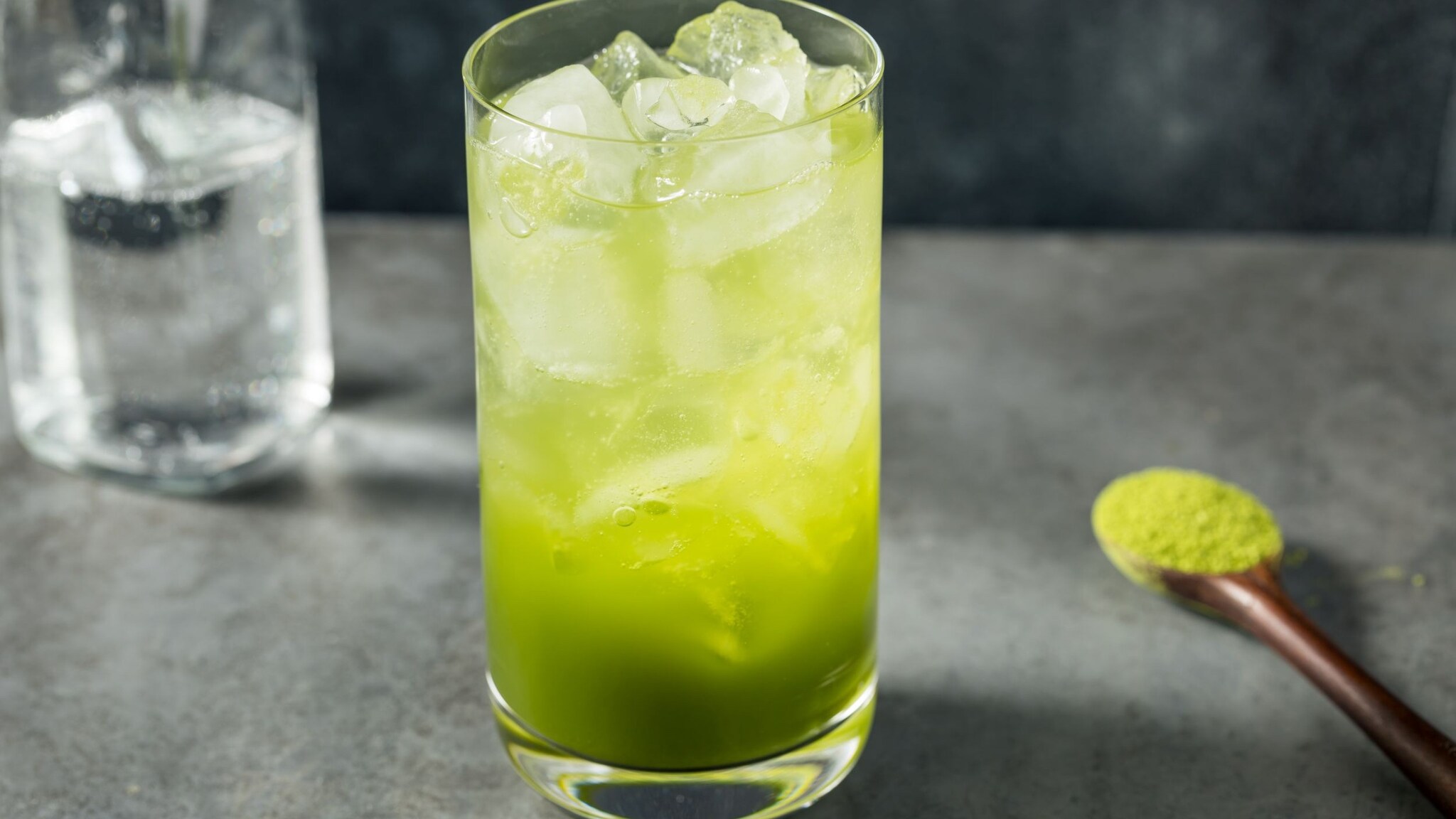 Glas mit grünem Matcha-Getränk und Eiswürfeln auf einer grauen Unterlage.